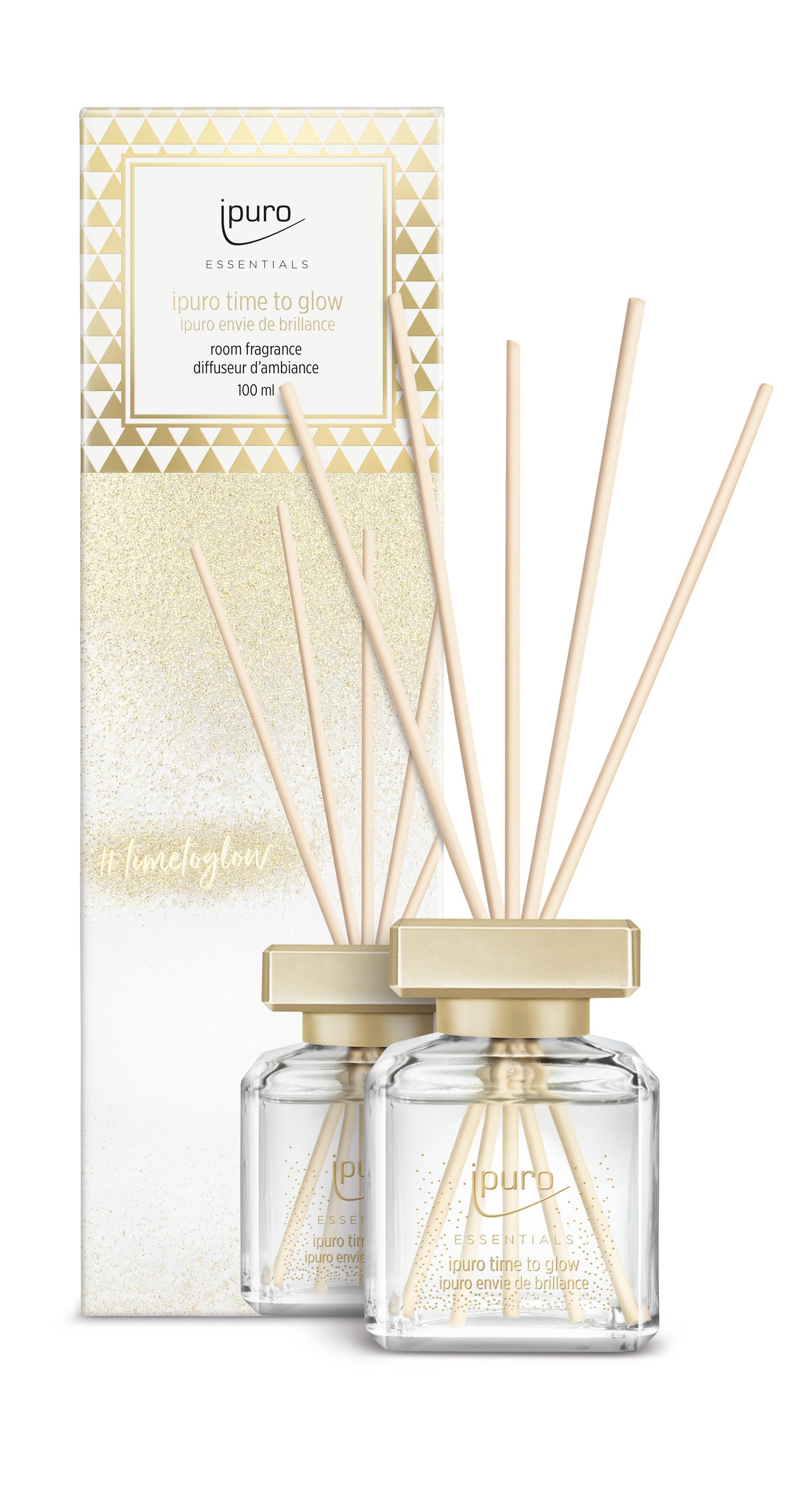 Der ESSENTIALS ipuro time to glow Raumduft von ipuro präsentiert sich in einem Glasflakon mit goldenem Deckel und Holzstäbchen mit orientalisch-floralen Noten wie Tonkabohne und Kaschmirholz vor einer glitzernden, geometrischen Verpackung.