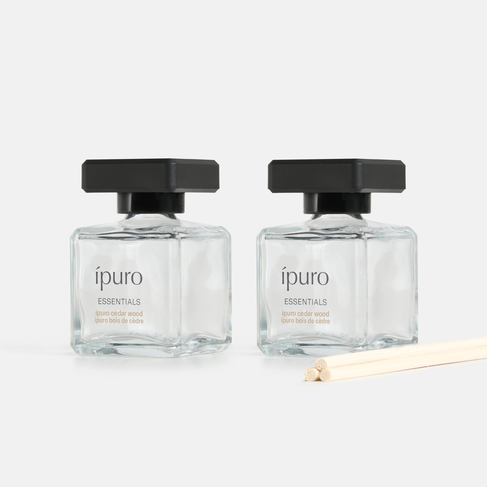 Zwei klare Glasflaschen mit schwarzen Verschlüssen, beschriftet mit "ipuro cedar wood" von ipuro, stehen nebeneinander auf einer weißen Fläche. Drei helle Schilfrohrstäbchen davor verströmen den frischen und beruhigenden Duft von Zedernholz.