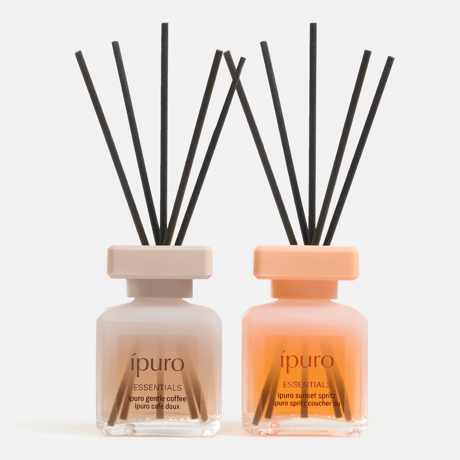 Zwei gläserne Diffusorflaschen von ipuro mit den Bezeichnungen "sunset spritz" und "gentle coffee" stehen mit quadratischen Kappen und schwarzen Stäbchen nebeneinander - ideal für einen sommerlichen Duft. Teil des Geschenk-Sets von ipuro.