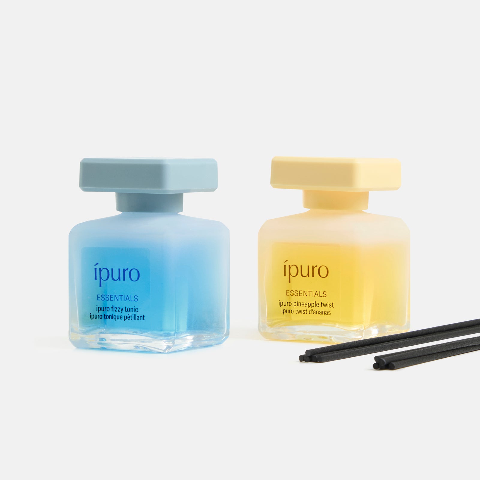 Das ipuro ESSENTIALS Ananas Twist & Fizzy Tonic Geschenk-Set besteht aus zwei quadratischen Glasdüsen mit passenden Kappen und bietet Zitrusnoten und tropischen Duft. Das Set enthält drei schwarze Stäbchen mit dem Logo von ipuro auf weißem Hintergrund.