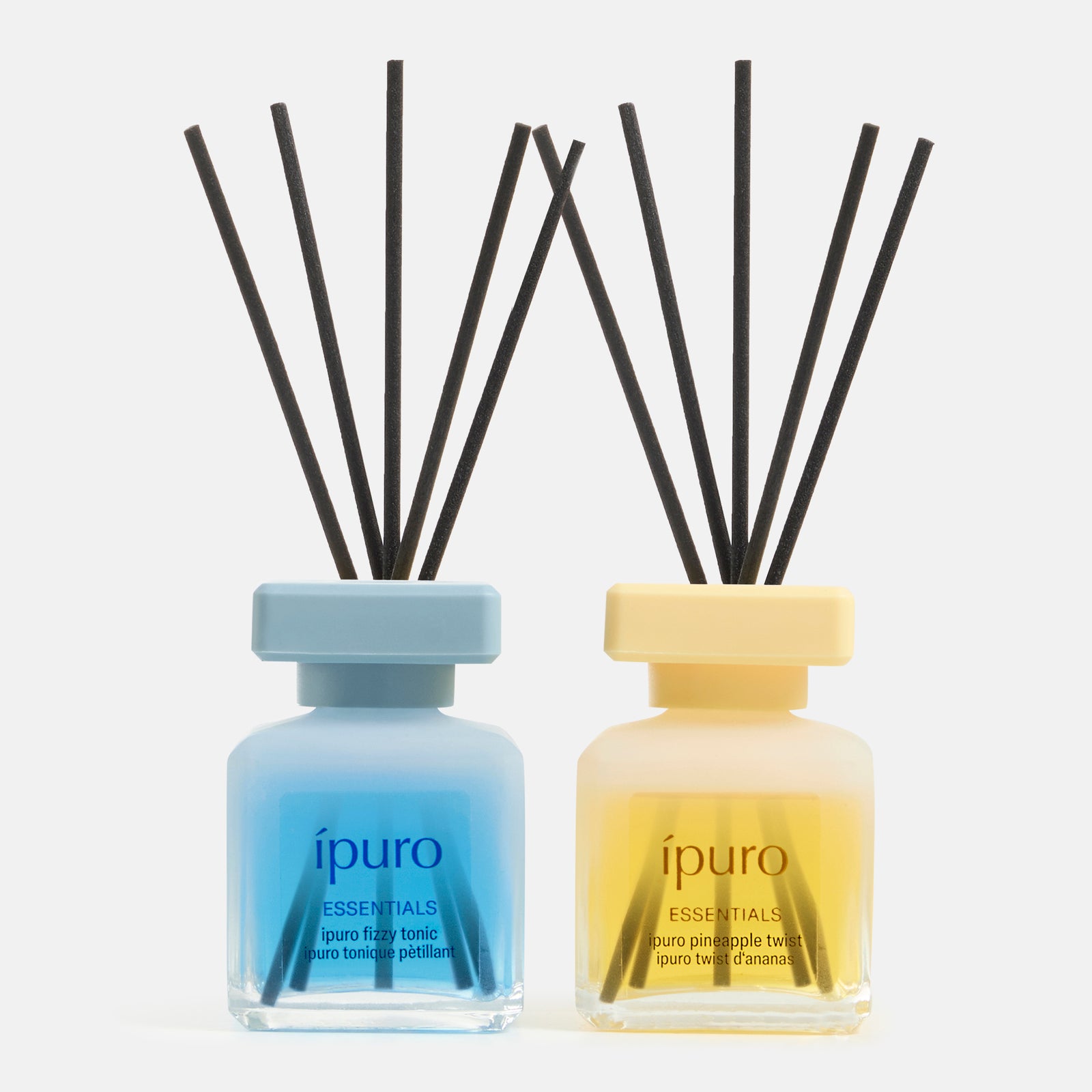 Das Geschenk-Set ipuro pineapple twist & fizzy tonic besteht aus zwei Glasdiffusoren von ipuro in den Farben blau und gelb mit schwarzen Schilfhalmen, die lebendige Düfte auf einem klaren weißen Hintergrund bieten.