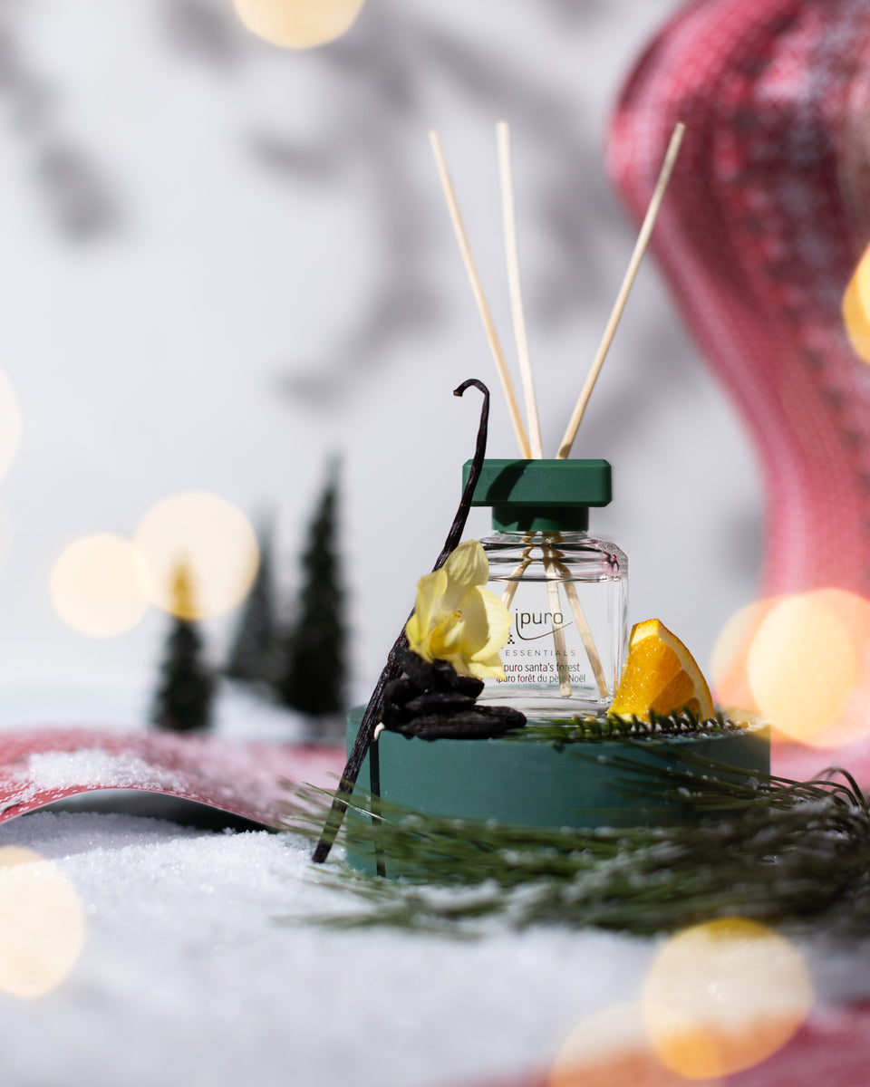 Eine klare Flasche ipuro ESSENTIALS Raumduft santa's forest diffuser mit Holzstäbchen steht auf einem grünen Sockel, umgeben von Tannennadeln, Vanilleschoten, gelben Blüten und Orangenscheiben inmitten von Schnee und Miniaturbäumen für eine festliche Winterszene.