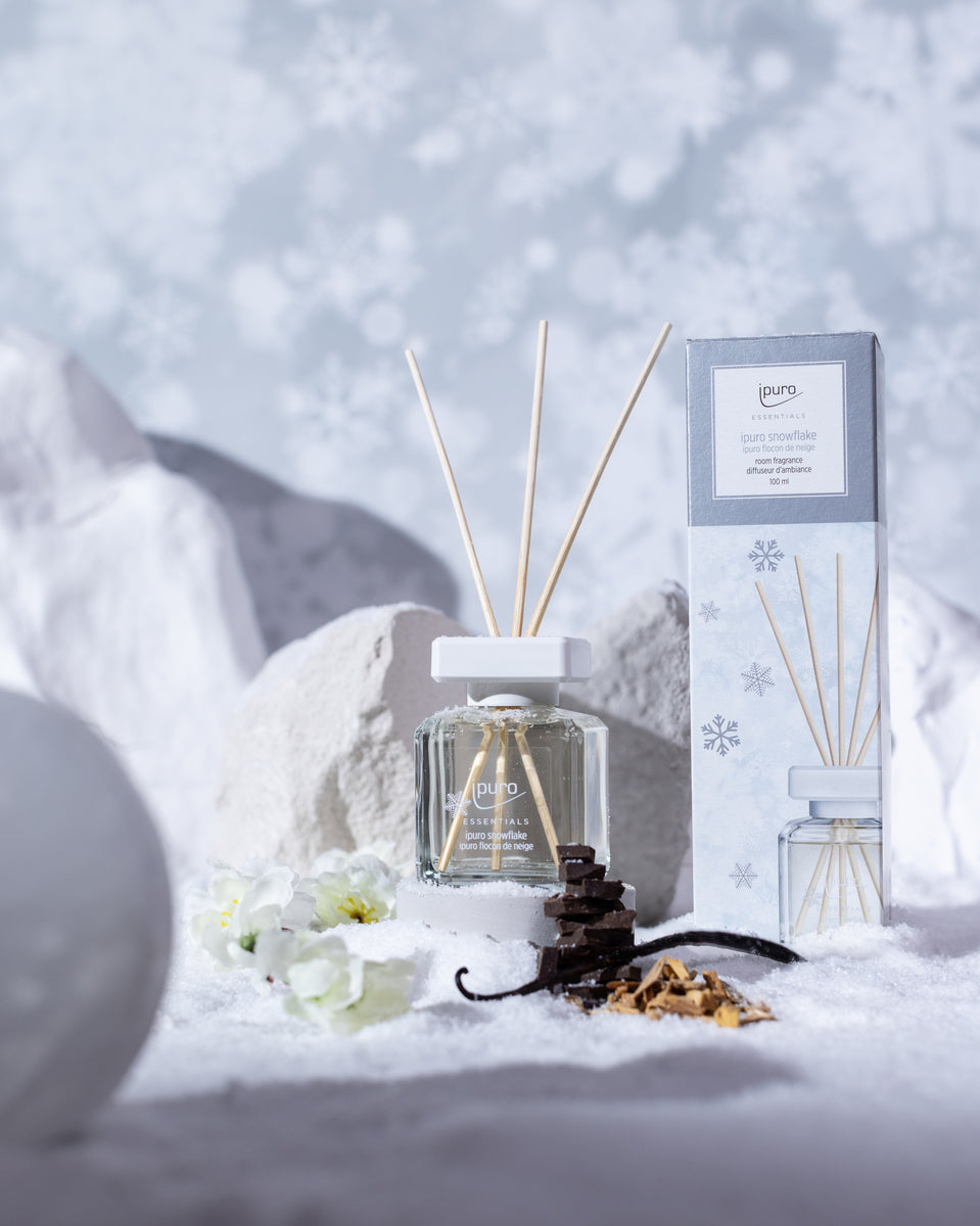 Ein ESSENTIALS Raumduft Snowflake Diffusor von ipuro mit Holzstäben sitzt auf Schnee neben seiner Box, umgeben von weißen Blumen und getrockneten Pflanzen. Mandelblüten-Noten und hellgraue Felsen vervollständigen die winterliche Szene.