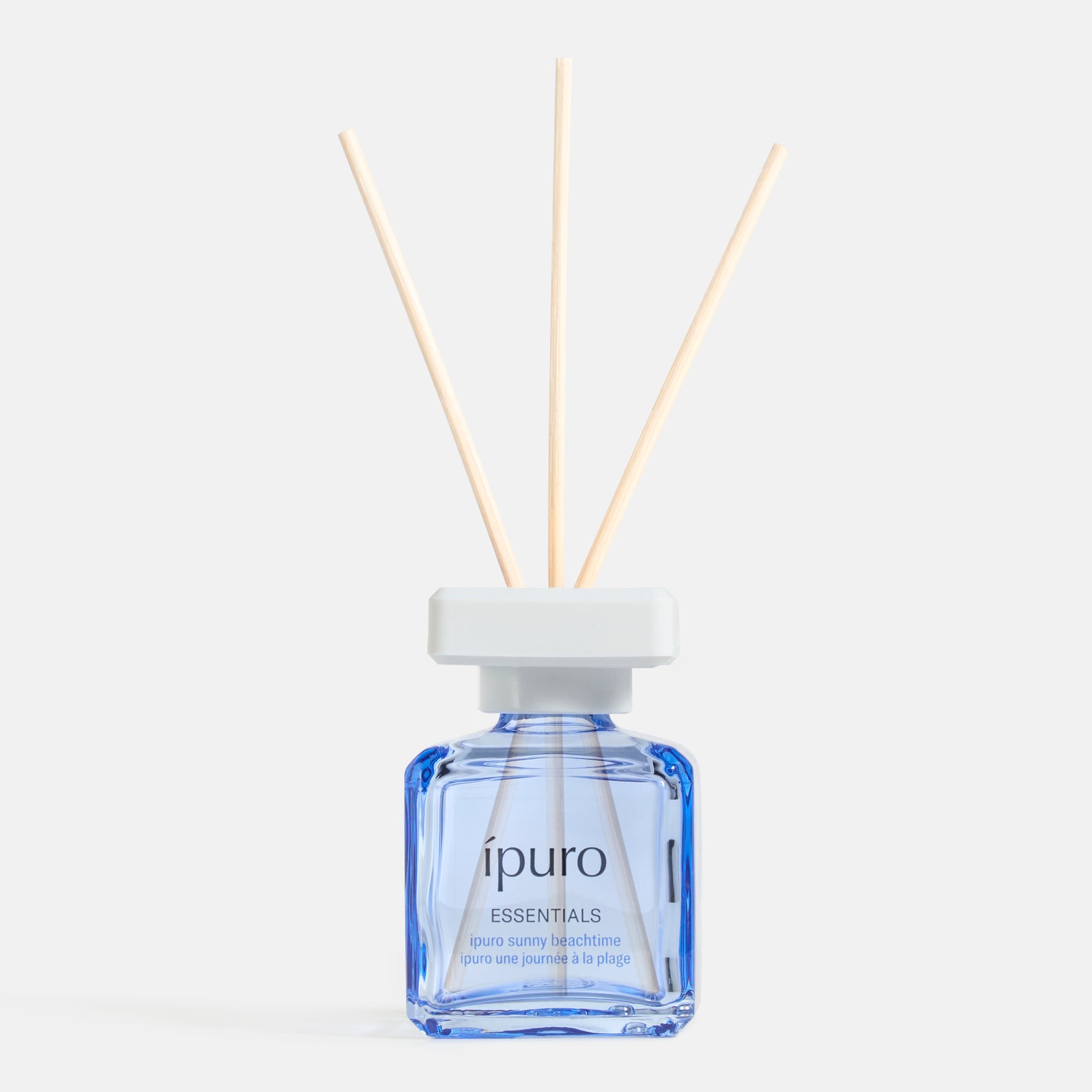 Ein blauer Glasflakon von ipuro sunny beachtime mit weißer Kappe und drei hölzernen Diffusorstäbchen erinnert an Sommer und Sonne vor einem schlichten weißen Hintergrund.