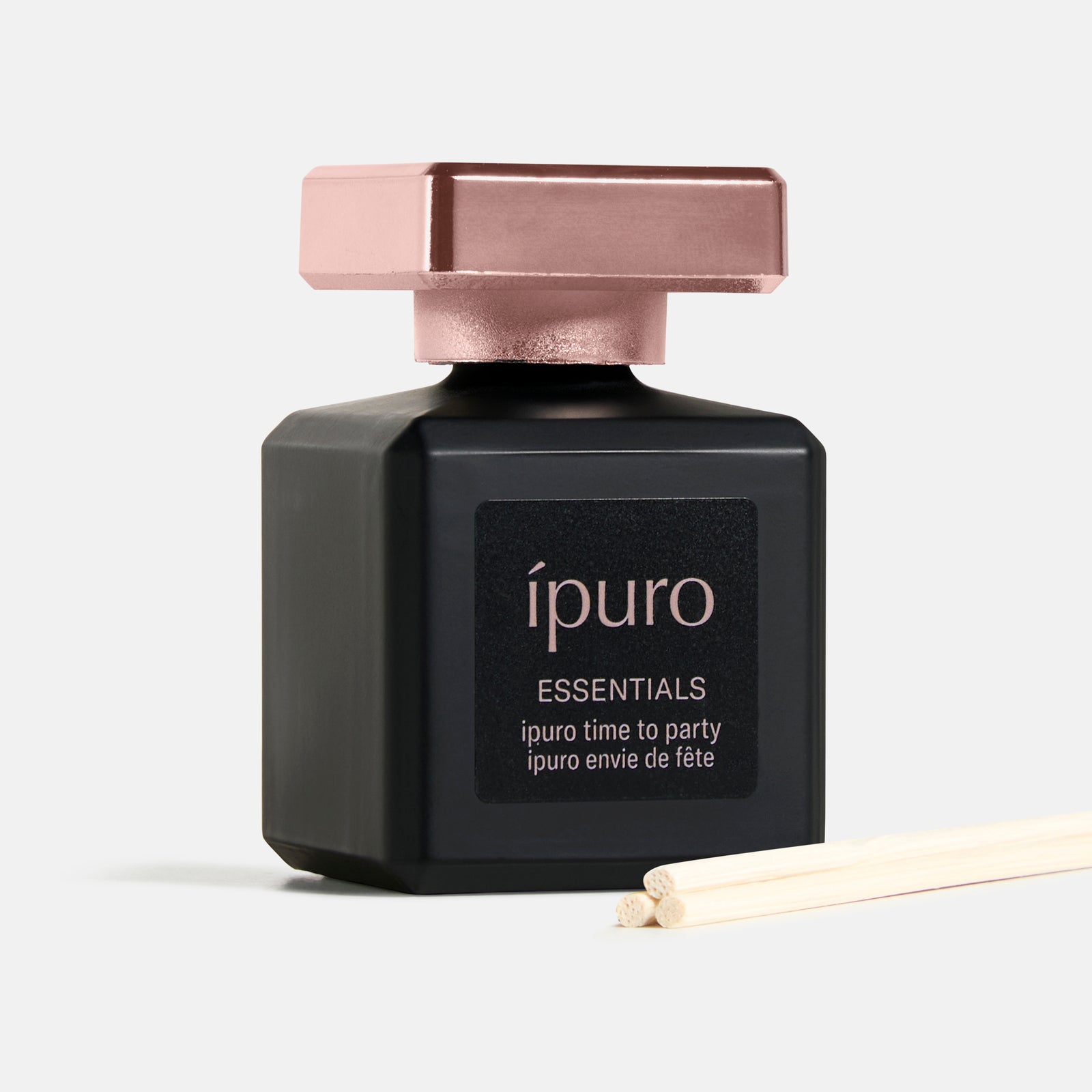 Ein schwarzer Flakon des Duftes ipuro Raumduft time to party mit roségoldener Kappe und Etikett wird neben zwei hölzernen Diffusorstäben auf einem hellen Hintergrund präsentiert.