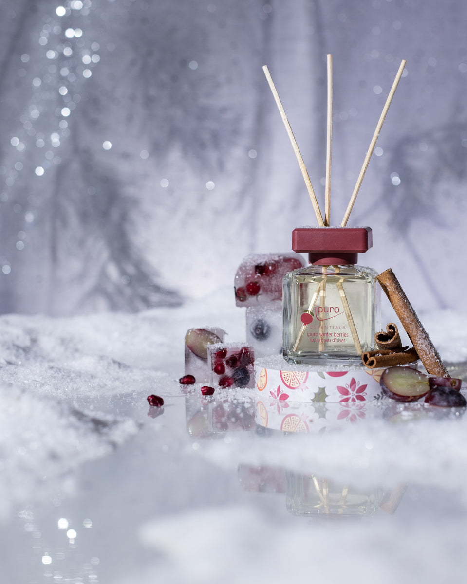 Das ipuro ESSENTIALS Raumduft-Set Snowflake & Winter Berries zeigt einen Glasdiffusor mit Holzstäbchen auf Schnee, umgeben von Granatapfelkernen, Fruchtscheiben, Zimtstange und einer dekorativen Box vor einer glitzernden Winterkulisse.