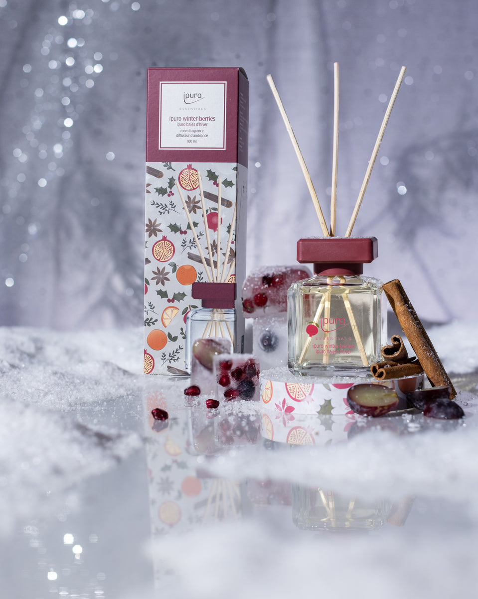 Der ipuro ESSENTIALS Raumduft Winter Berries, ein Glasrohrdiffusor mit kastanienbrauner Kappe und drei Stäbchen, sitzt in seiner dekorierten Schachtel inmitten von Kunstschnee, Zimtstangen und Beeren für eine festliche Winterszene mit warmen Beeren- und Nelkennoten.
