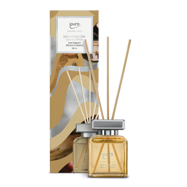 Ein Flakon des Raumdufts ipuro Essentials Winter Cider von ipuro mit hellbrauner Flüssigkeit, silbernem Verschluss und Rohrdiffusorstäbchen neben der beige-goldenen dekorativen Verpackung.