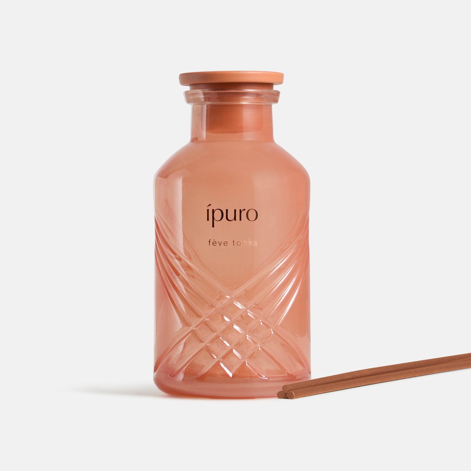 Ein rosafarbener ipuro Raumduft-Fève Tonka-Glasflakon mit passender Kappe und braunem Schilfrohr, abgebildet auf weißem Hintergrund, erinnert an einladende Düfte von Tonkabohne und Vanille.