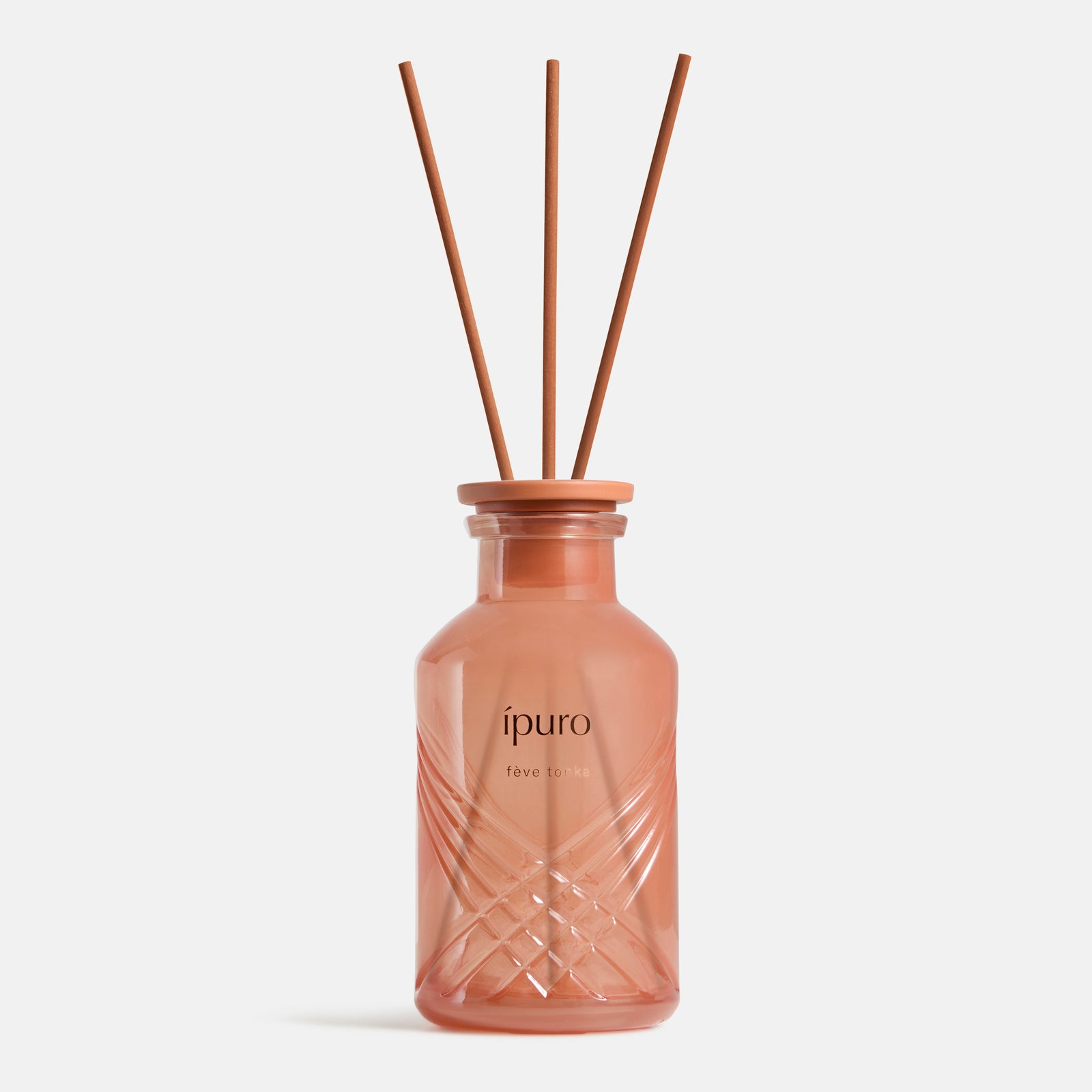 Ein rosafarbener Milchglasflakon mit geometrischen Mustern und der Aufschrift "ipuro fève tonka" von ipuro enthält drei braune Schilfrohrstäbchen mit den Noten Vanille und Tonkabohne. Er ist vor einem schlichten weißen Hintergrund fotografiert.