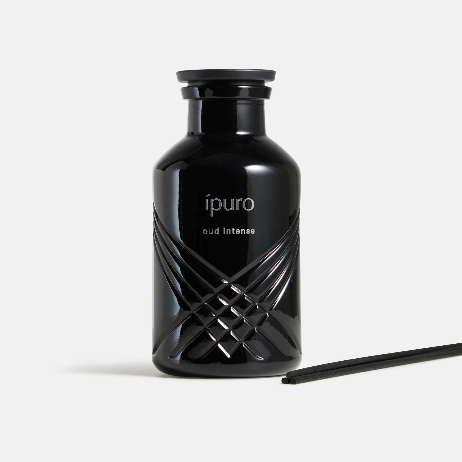 Ein schwarzer, glänzender Flakon von ipuro Raumduft oud intense, der mit dem Markennamen beschriftet ist, weist in der Nähe des Bodens ein strukturiertes Kreuzmuster auf. Daneben liegt ein schwarzes Schilfrohr, das auf die Düfte von Oud und Safran hinweist. Der Hintergrund ist einfarbig weiß.