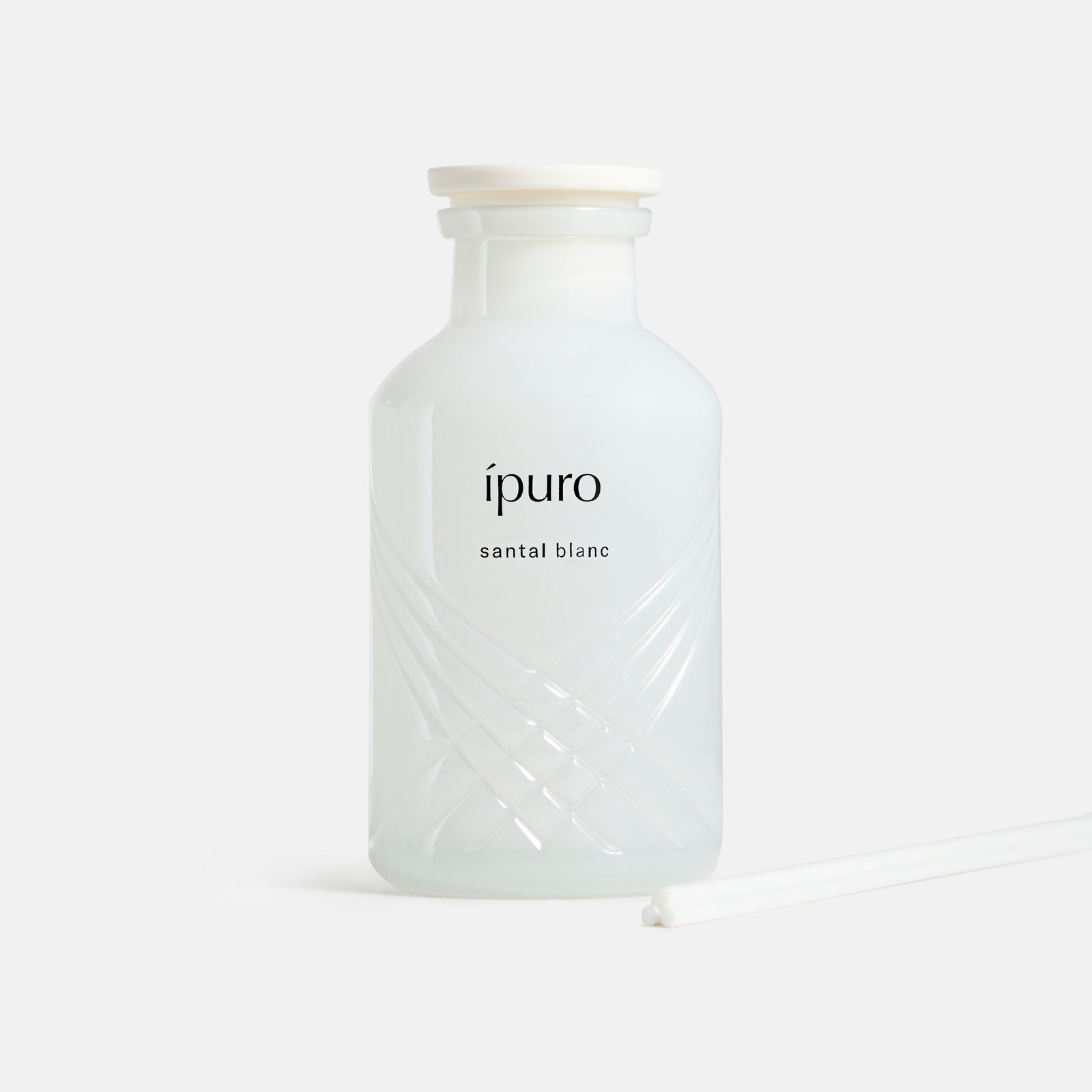 Eine Flasche aus weißem Milchglas mit weißer Kappe und der Aufschrift ipuro santal blanc Raumduft von ipuro weist ein subtiles Kreuzmuster auf. Sie steht neben einem weißen Schilfrohr, das vor einem schlichten weißen Hintergrund steht.