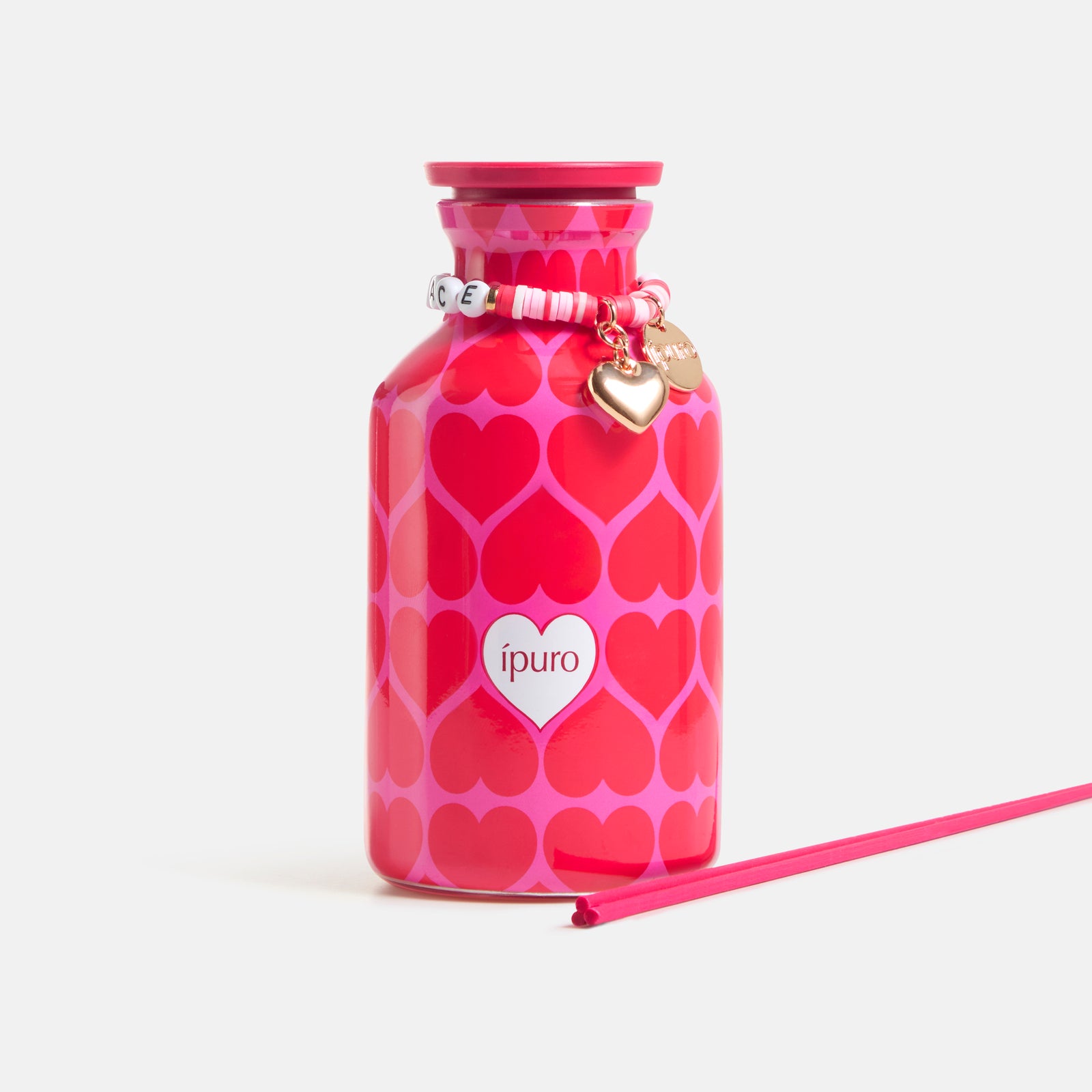 Die LIMITED EDITION ipuro love's embrace Raumduft 240ml von ipuro besteht aus einer rosafarbenen Flasche mit sich überlappenden Herzen, einem herzförmigen Anhänger und einem Perlenarmband. Auf der Vorderseite befindet sich der Schriftzug "ipuro". Daneben liegt ein rosafarbener Stab auf weißem Hintergrund.