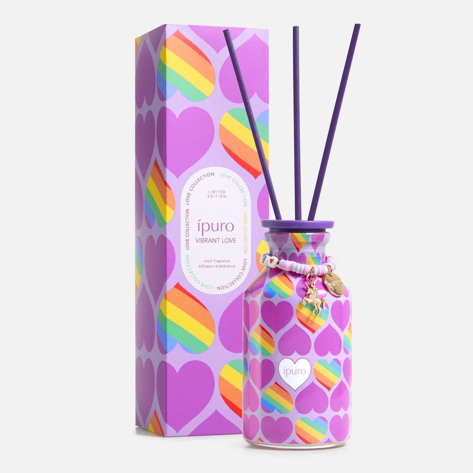 Ein lilafarbener Diffusor-Flakon mit Regenbogenherz-Muster und lilafarbenem Schilfrohr steht neben einer passenden Box. Der LIMITED EDITION ipuro vibrant love Raumduft 240ml von ipuro enthält fruchtige Noten und Maiglöckchen sowie dekorative Anhänger.