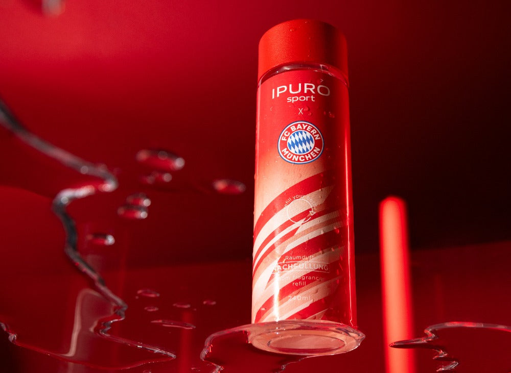 Eine rote Flasche mit der Aufschrift IPURO sport und dem Logo des FC Bayern München steht aufrecht auf einer spiegelnden Fläche mit Wassertropfen vor einem roten Hintergrund.