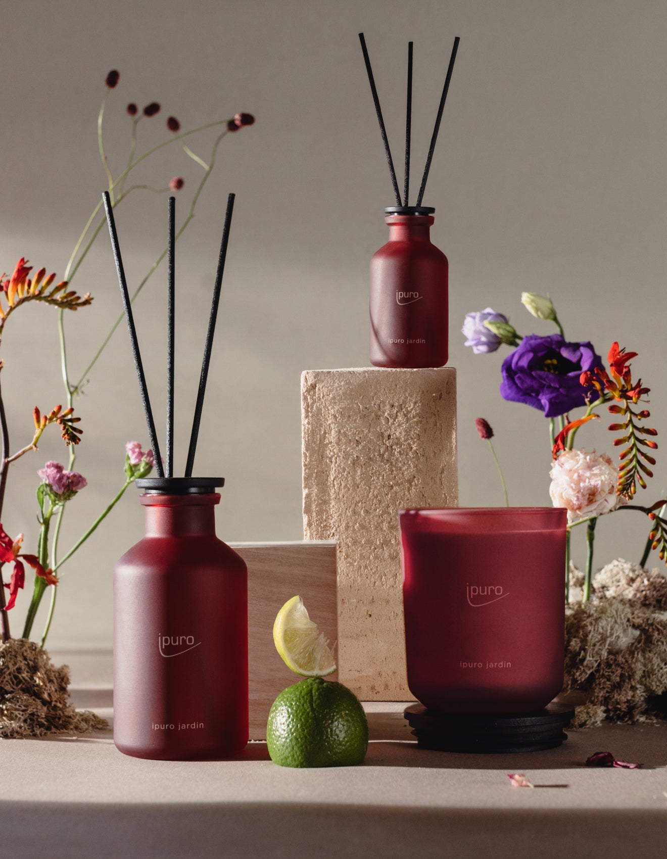 Zwei rote Glasduftdiffusoren und eine passende Kerze werden mit schwarzem Schilfrohr in Szene gesetzt, umgeben von Blumen, einer Limette und einem Steinblock, wodurch ein elegantes und natürliches Arrangement entsteht.