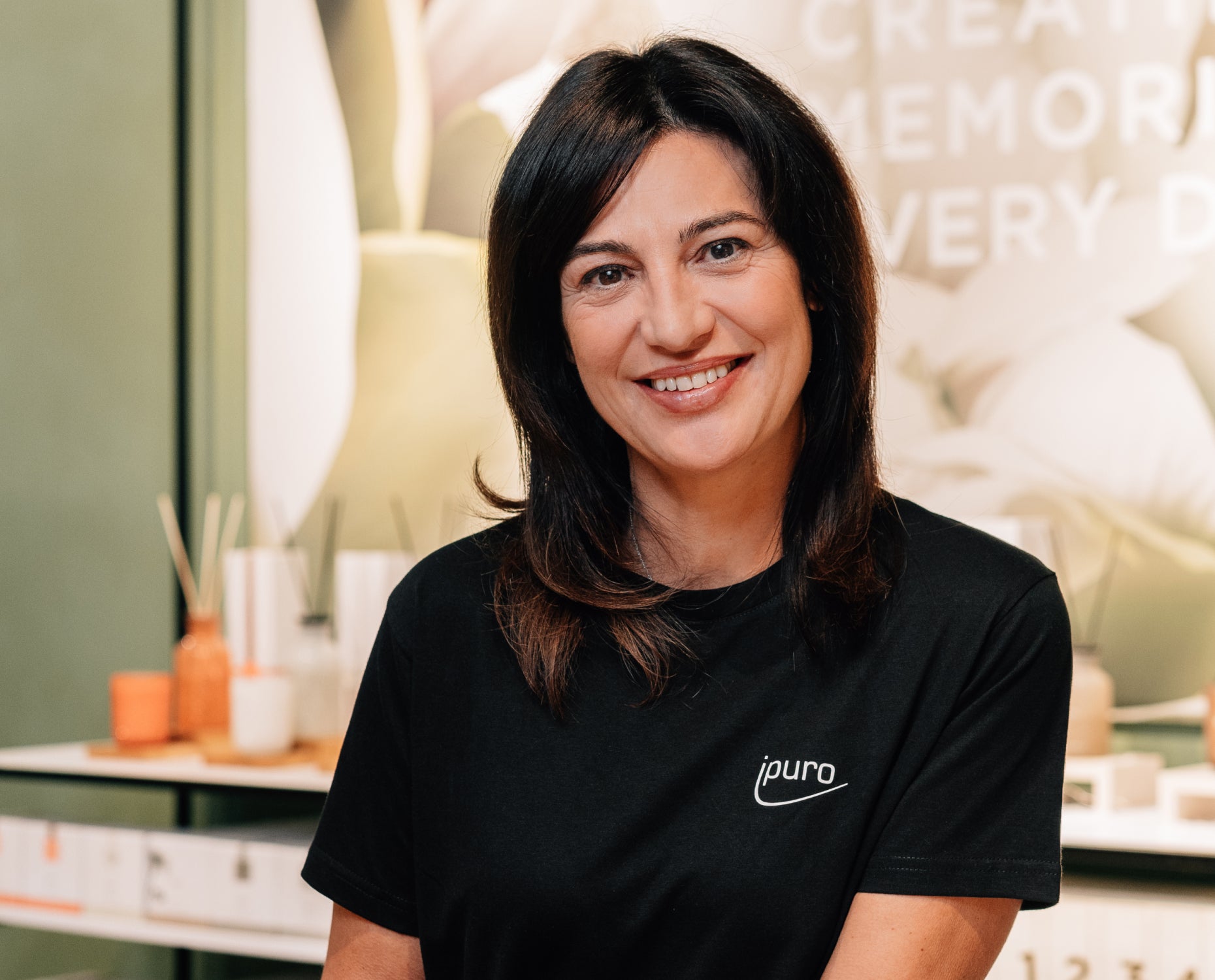Eine Frau mit dunklem Haar lächelt, trägt ein schwarzes Ipuro-Shirt und steht in einem Innenraum vor Regalen mit Diffusoren und einem verschwommenen Schild im Hintergrund.