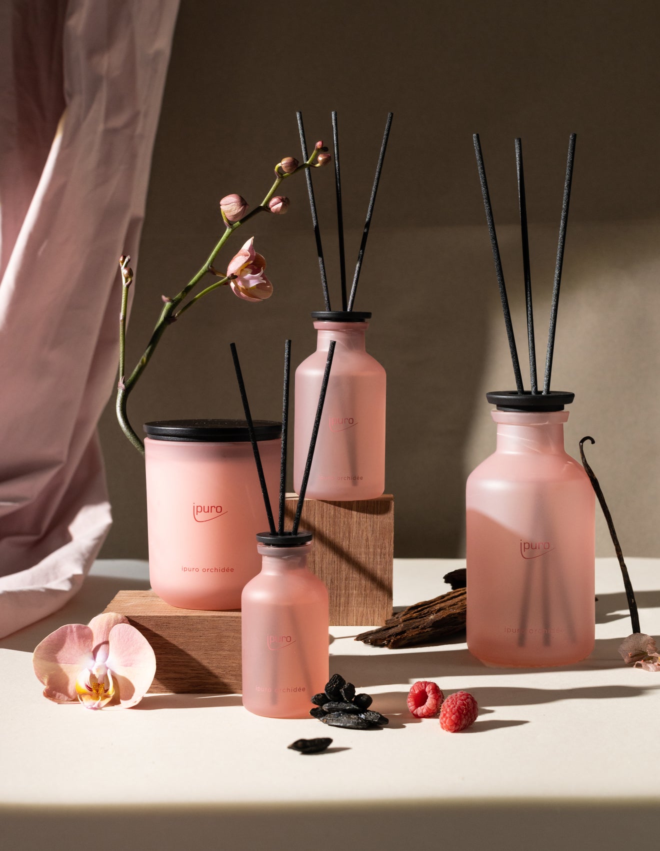 Vier rosafarbene Duftzerstäuber aus Milchglas und eine Kerze sind mit schwarzem Schilf, Himbeeren, Vanilleschoten und Orchideen auf einer beigen Fläche arrangiert, während im Hintergrund ein rosafarbener Vorhang drapiert ist.