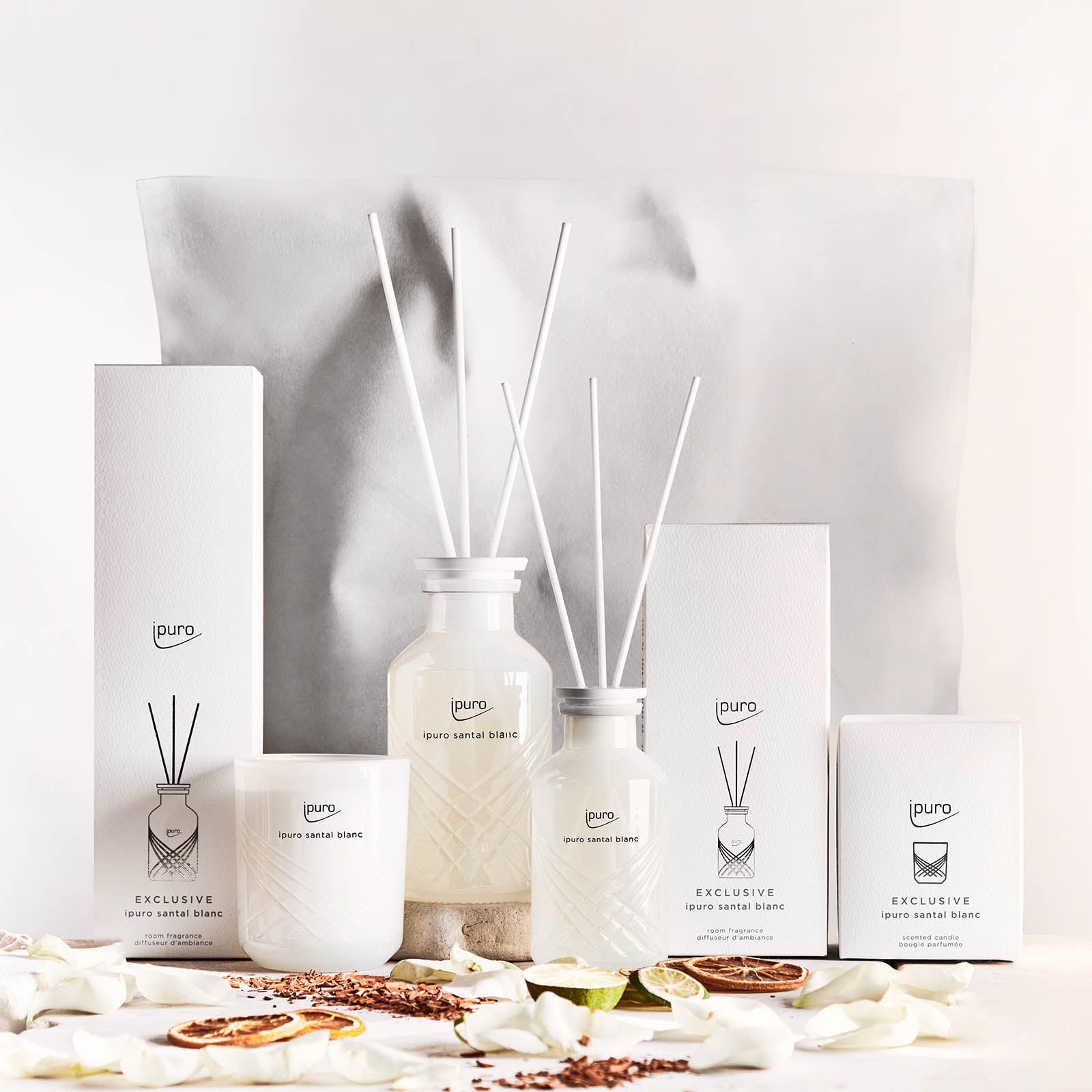 Ein Display mit EXCLUSIVE ipuro santal blanc Raumduft von ipuro, mit Düften von Sandelholz und weißer Orchidee in einer minimalistischen weißen Verpackung, umgeben von Trockenblumen und Potpourri vor einem neutralen Hintergrund.