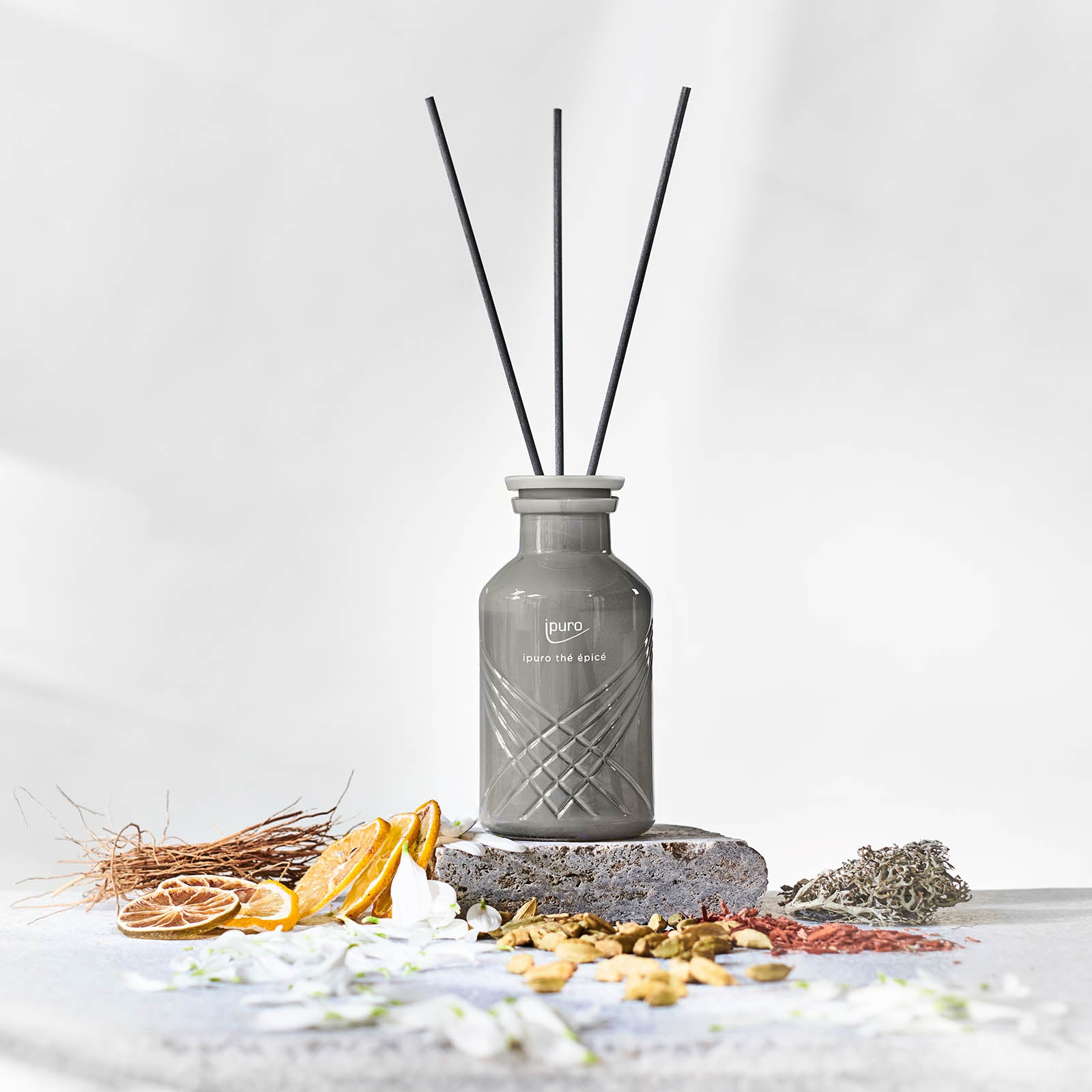 Der EXCLUSIVE ipuro thé epicé Raumduft von ipuro besteht aus einem grauen Glasdiffusor mit schwarzem Schilf, umgeben von getrockneten Orangenscheiben, Zweigen, weißen Blüten und Pflanzen; Noten von Matcha-Tee und Zitrone sorgen für ein erfrischendes Aroma.