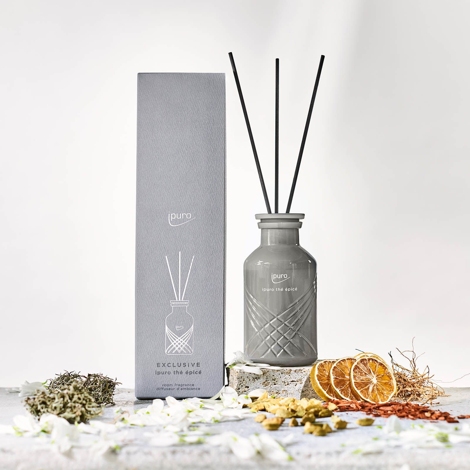 Das EXCLUSIVE ipuro thé epicé Raumduft von ipuro mit grauem Diffusor, drei schwarzen Stäben und passender Schachtel wird zwischen Trockenblumen und Orangenscheiben auf einer hellen Fläche präsentiert und sorgt für einen hellen, minimalistischen Look.