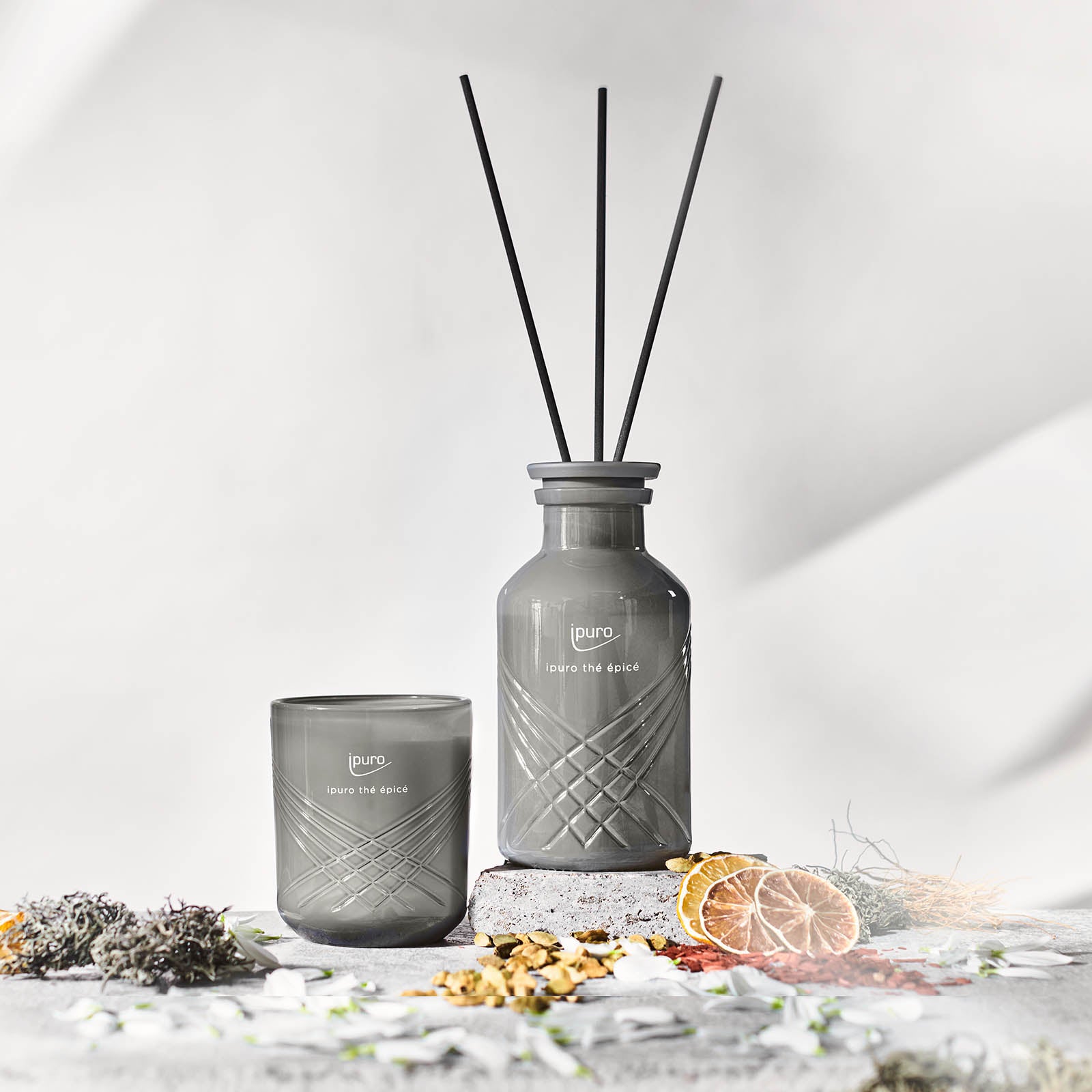 Die graue EXCLUSIVE ipuro thé epicé Duftkerze und der dazu passende Diffusor mit schwarzem Schilfrohr enthalten Noten von Sandelholz und Matcha-Tee, die auf einer strukturierten Oberfläche mit getrockneten Pflanzen dargestellt sind und das charakteristische Aroma von ipuro verkörpern.