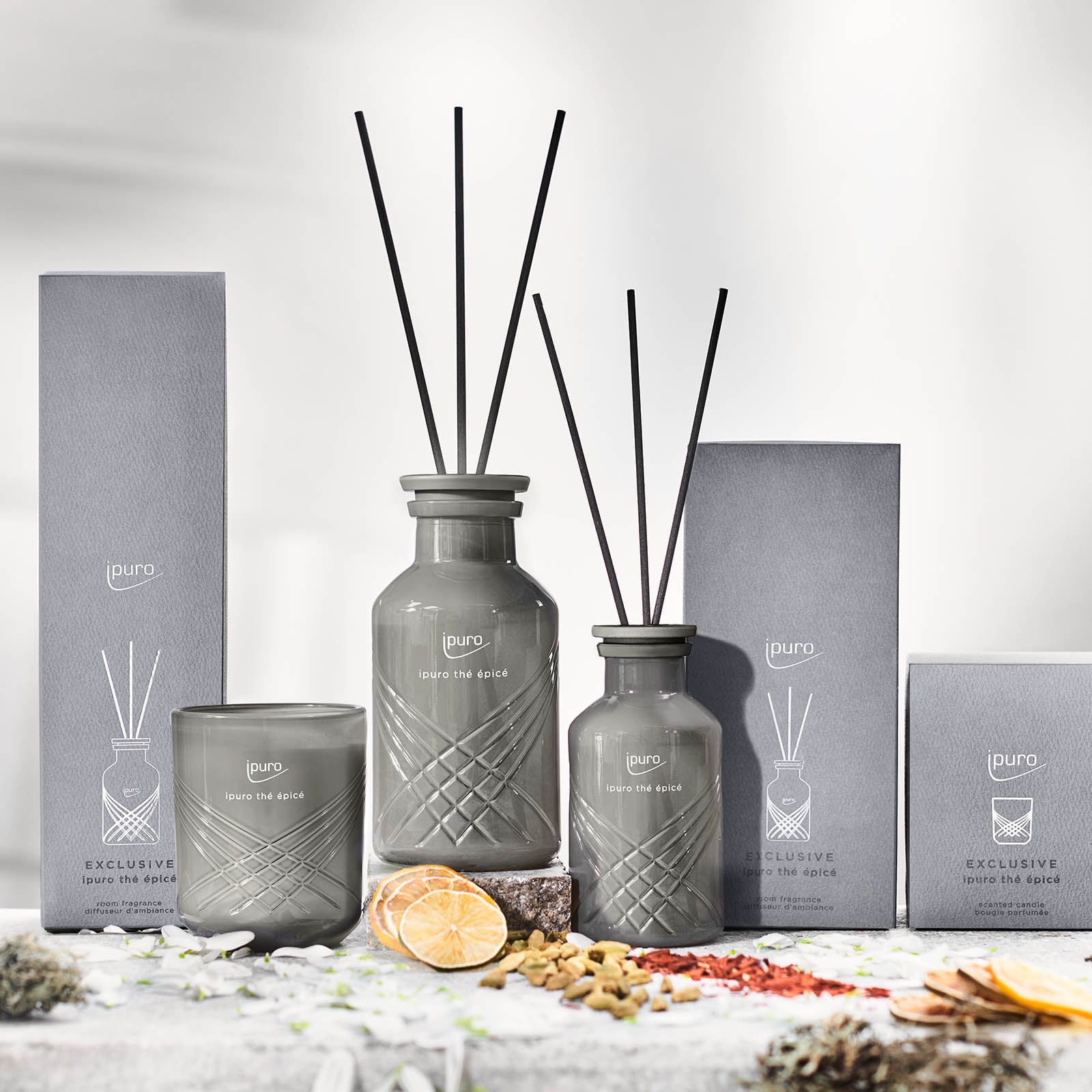 Die EXCLUSIVE ipuro thé epicé Duftkerze von ipuro und die dazu passenden Diffusoren mit schwarzen Stäbchen sitzen fein säuberlich neben den grauen Boxen. Getrocknete Zitronenscheiben, verstreute Gewürze und ein Hauch von Jasmin zieren die weiße Fläche davor.