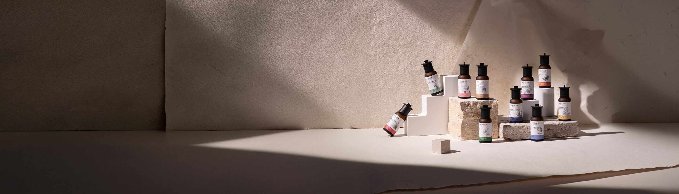 Eine Gruppe von Hautpflegeserumflaschen, die auf strukturierten Steinblöcken angeordnet sind, mit weichem Licht und Schatten vor einem beigen Papierhintergrund. Die Flaschen haben unterschiedliche Etiketten und sind ordentlich ausgestellt.