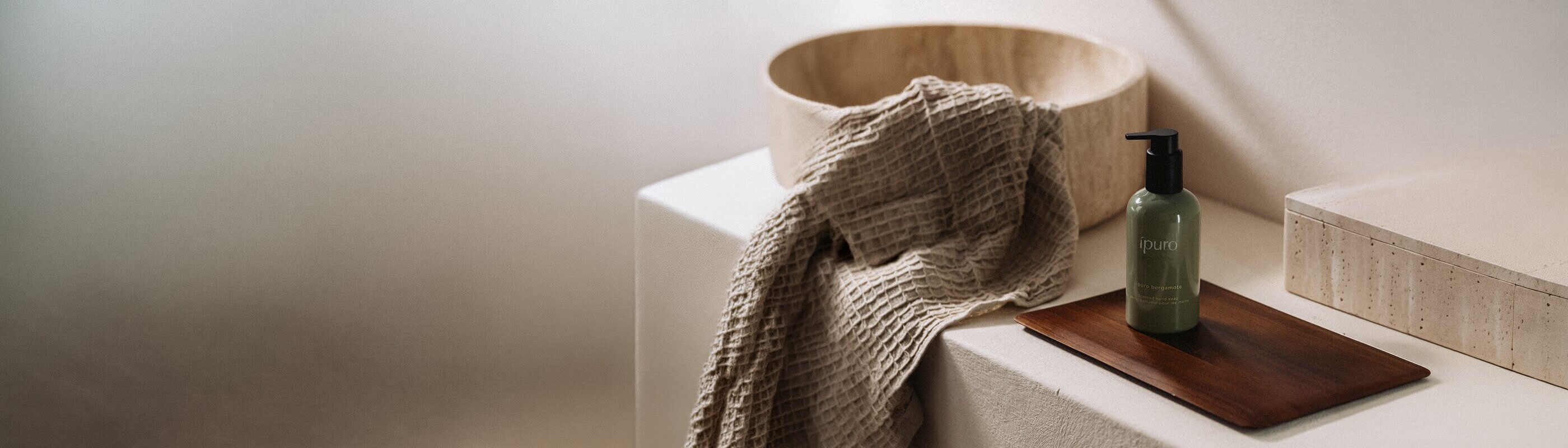 Eine braune Glasflasche mit der Aufschrift "Lavendel" und Schilfrohr-Diffusionsstäbchen, umgeben von Lavendelzweigen und grünen Blättern, steht auf einer hellen Fläche vor einem weichen, verschwommenen Hintergrund.