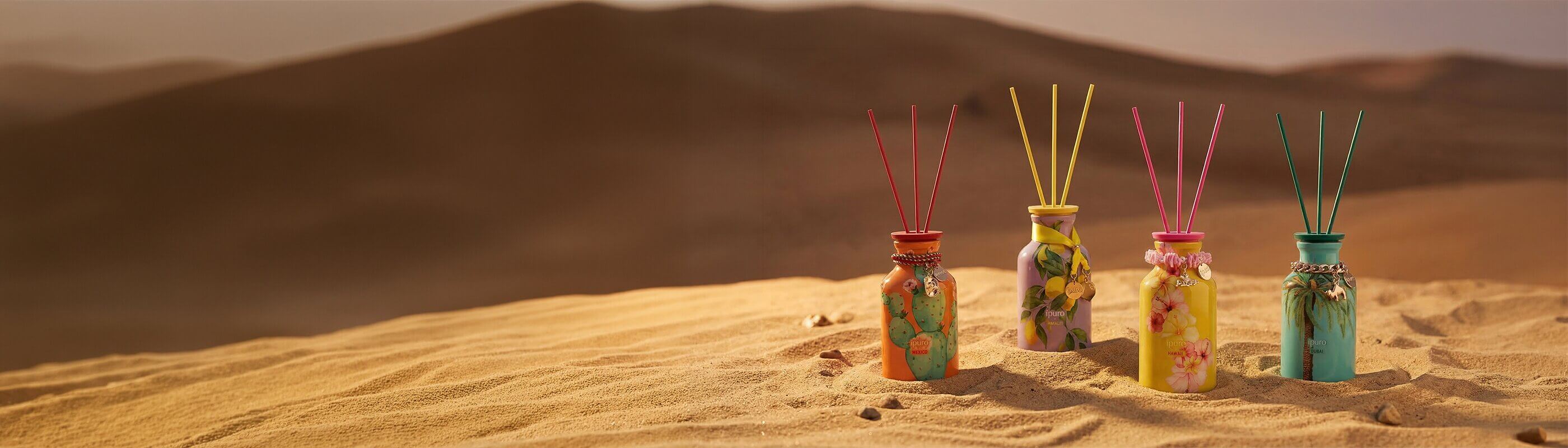 Vier farbenfrohe, dekorative Schilfrohrdiffusoren sind auf sandigem Wüstenboden platziert, wobei die Berge im Hintergrund in einem warmen, weichen Licht verschwimmen.