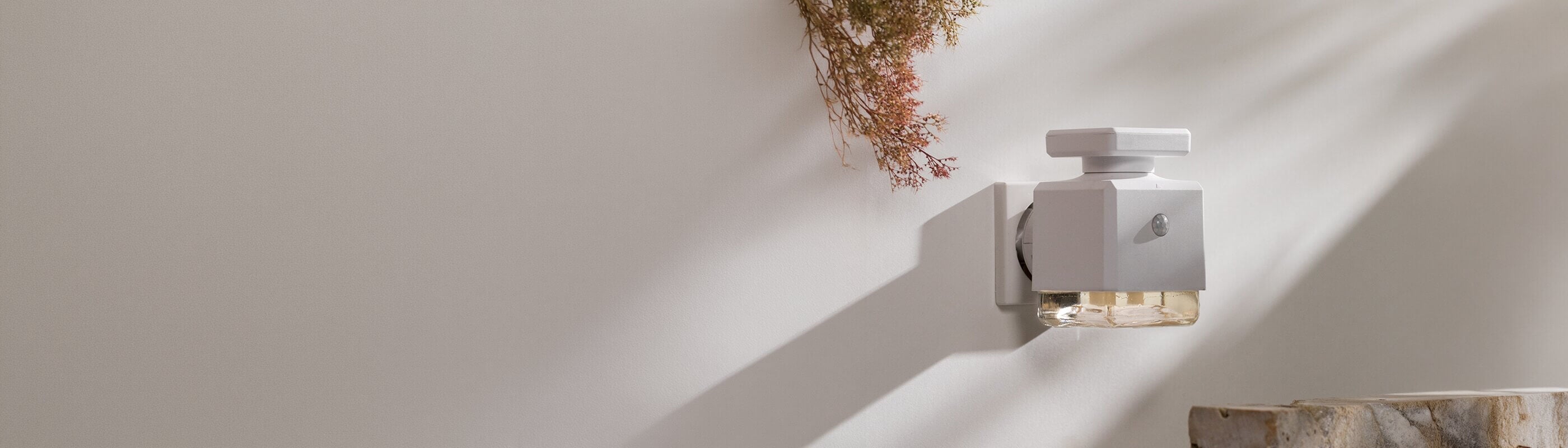 Ein weißer, an der Wand montierter Lufterfrischer mit einer Glasflasche, die einen Schatten auf eine helle Wand wirft, über der getrocknete Blumen hängen und unter der sich eine Steinfläche befindet.