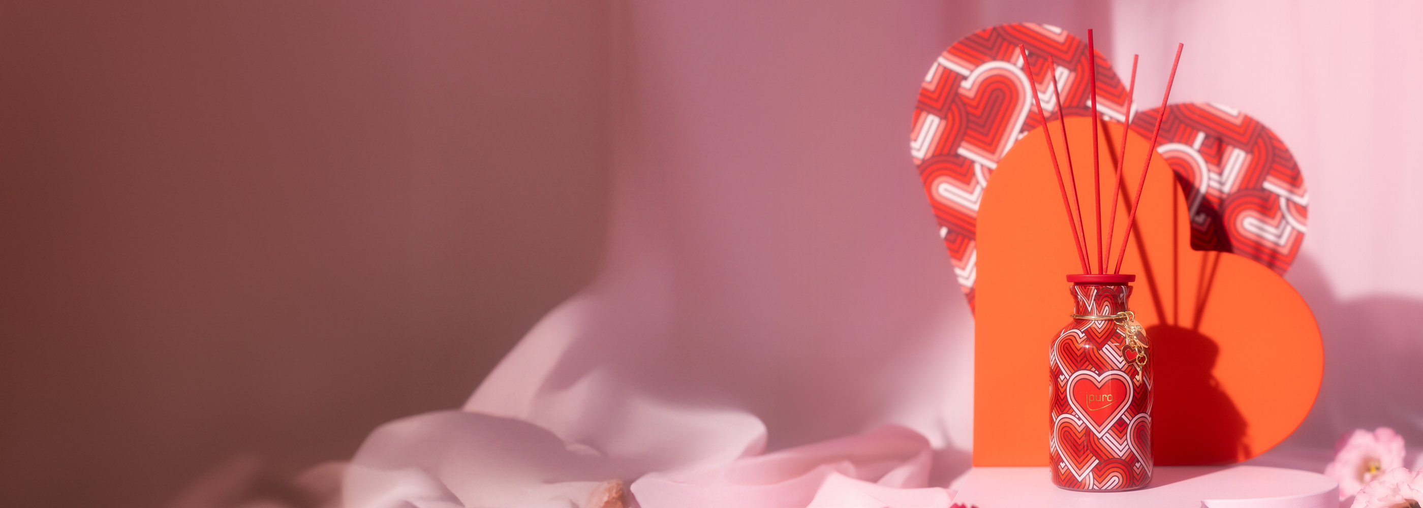 Bunte Diffusoren und Kerzen mit Herz- und LIEBE-Motiven sind auf einem rosa Stoffhintergrund ausgestellt, umgeben von Blumen, Nüssen und Himbeeren.