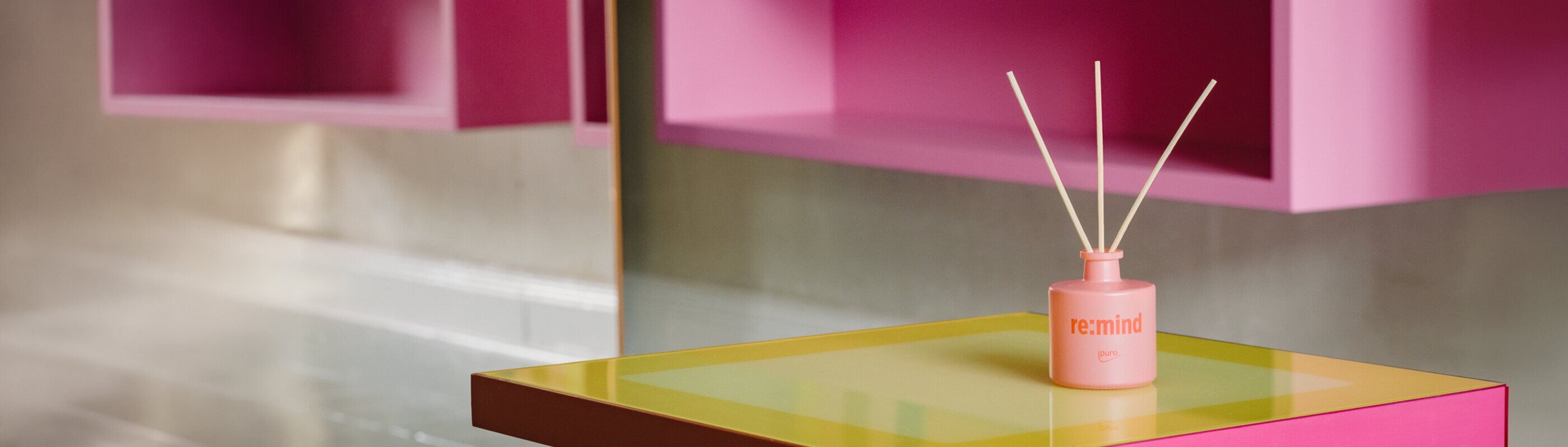 Ein rosafarbener Schilfrohrdiffusor mit der Aufschrift "remind" steht auf einem Tisch mit gelber Platte, im Hintergrund befinden sich rosafarbene geometrische Regale und daneben eine reflektierende Wandfläche.