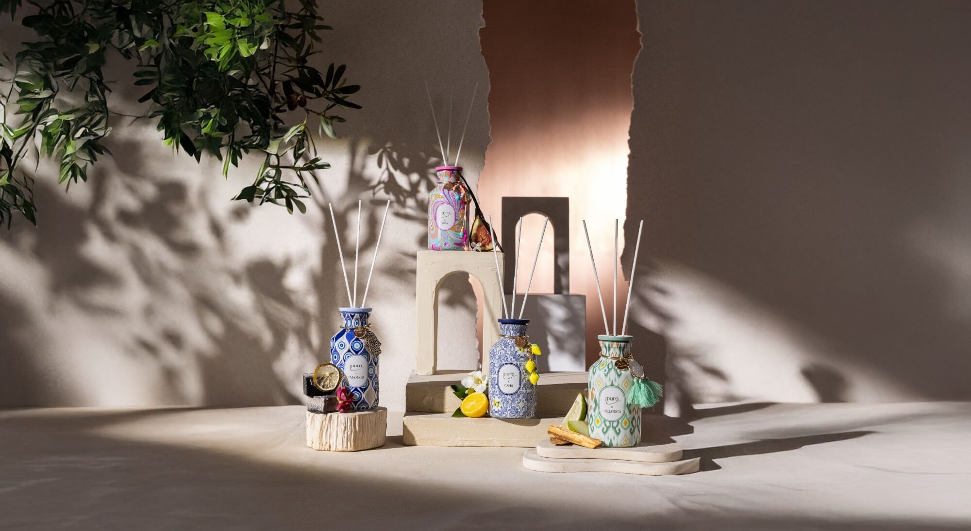 Vier dekorative Diffusoren in gemusterten Flaschen werden auf Podesten mit Früchten, Blumen und grünen Akzenten ausgestellt. Sonnenlicht und Schatten schaffen eine warme, heitere Atmosphäre vor einem neutralen Hintergrund.