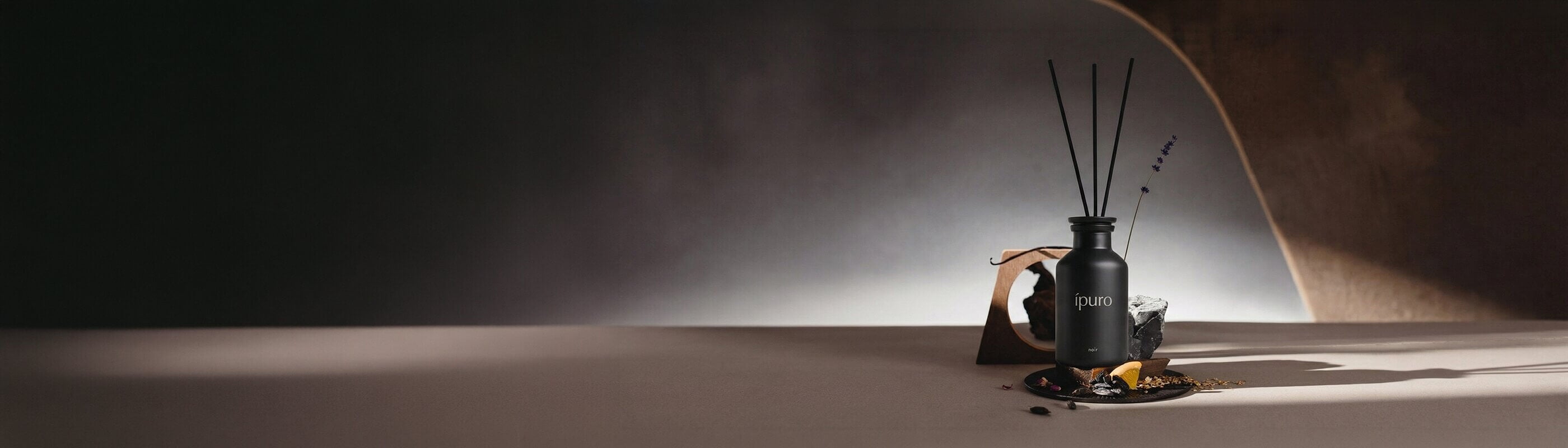 Ein schwarzer Ipuro noir-Schilfrohrdiffusor mit drei Stäben steht auf einem Autodach, mit verschwommenen Gebäuden und grünen Bäumen unter einem blauen Himmel im Hintergrund.