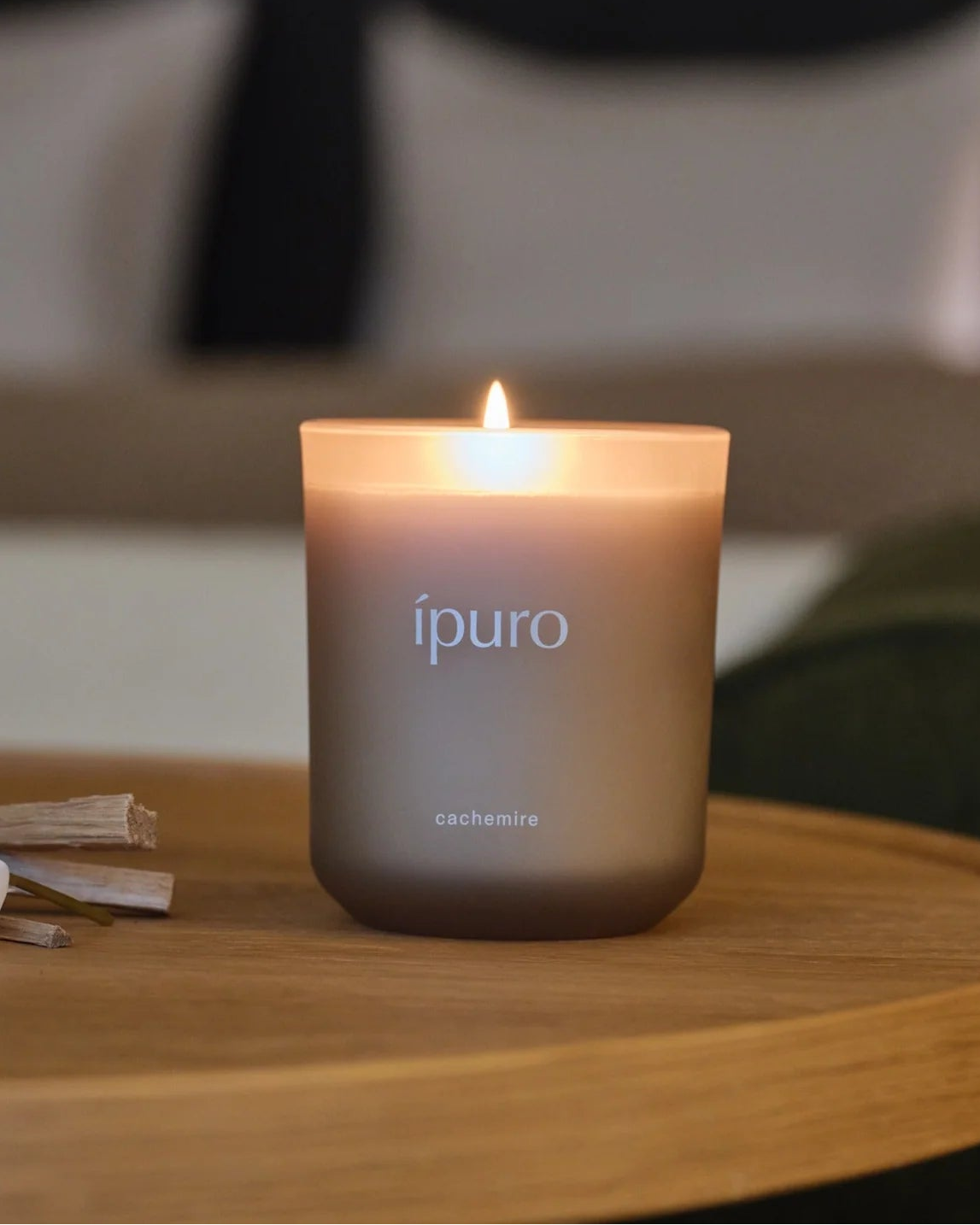 Eine brennende Duftkerze Ñpuro cachemireì in einem Milchglashalter steht auf einer hölzernen Fläche mit unscharfem Hintergrund.