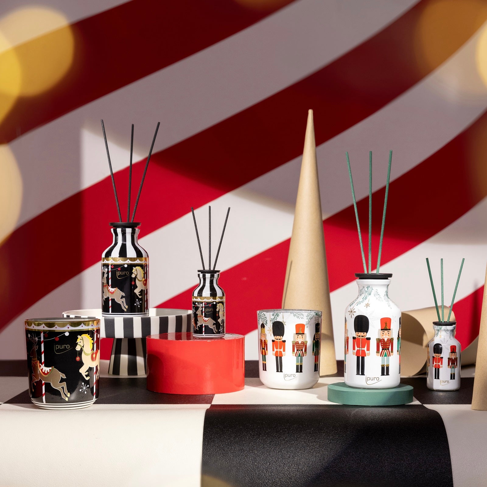 Das LIMITED EDITION ipuro finest season & magical market Geschenk-Set von ipuro zeigt Kerzen mit fruchtig-würzigem Duft und Zinnsoldaten-Motiven auf geometrischen Ständern vor einer leuchtend rot-weiß gestreiften Kulisse mit festlichen Lichtern.