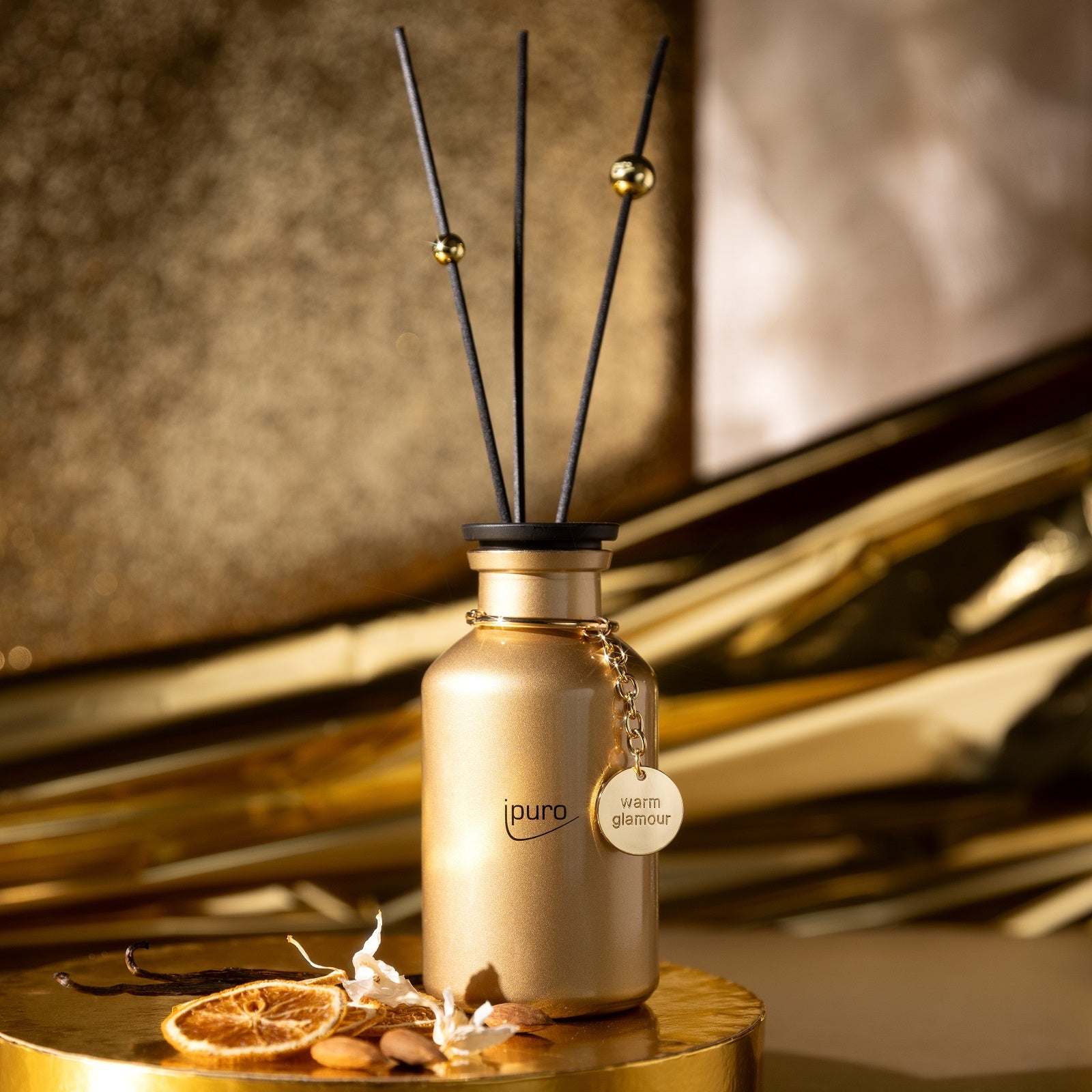 Die LIMITED EDITION ipuro warm glamour Raumduft von ipuro verfügt über einen goldenen Diffusor mit schwarzem Schilf und einem runden warm glamour Anhänger, der auf einer passenden Oberfläche mit getrockneten Orangenscheiben und goldenen Akzenten präsentiert wird - perfekt für jede winterliche Duftumgebung.