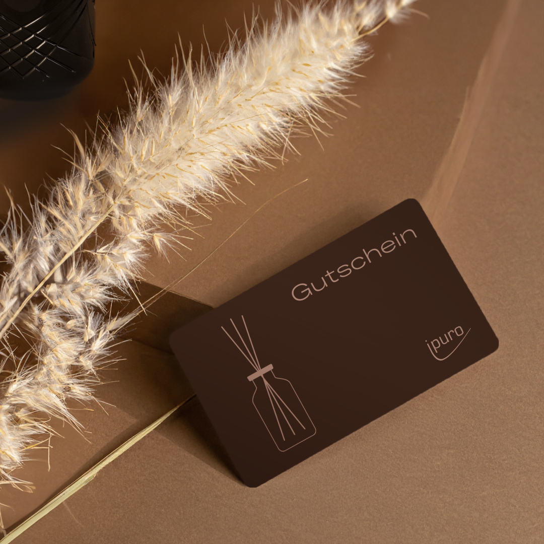 Eine ipuro-Gutschein-Geschenkkarte mit der Abbildung eines Schilfrohrdiffusors liegt auf einer braunen Fläche neben getrocknetem Pampasgras.