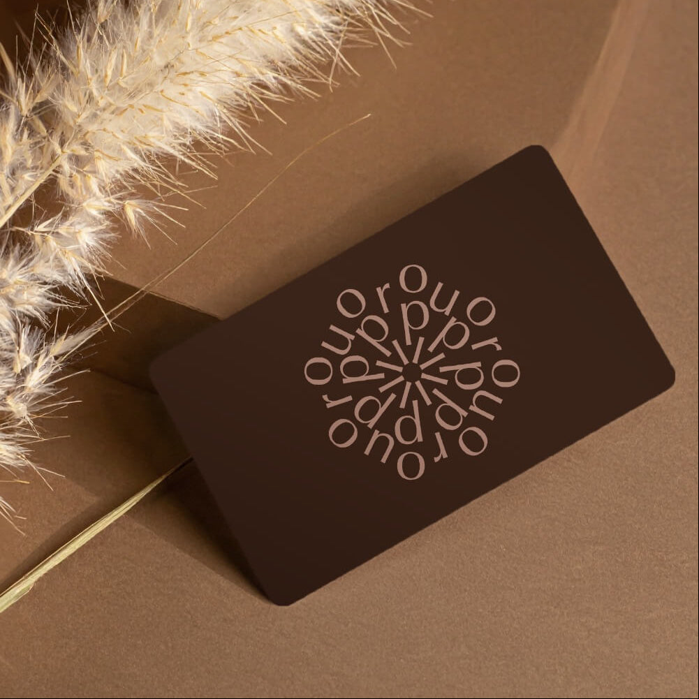 Eine braune Karte mit dem Wort "Ouro" in einem kreisförmigen Muster liegt auf einer braunen Fläche neben getrocknetem Pampasgras - ideal für ipuro Raumdüfte oder einen ipuro Gutschein.