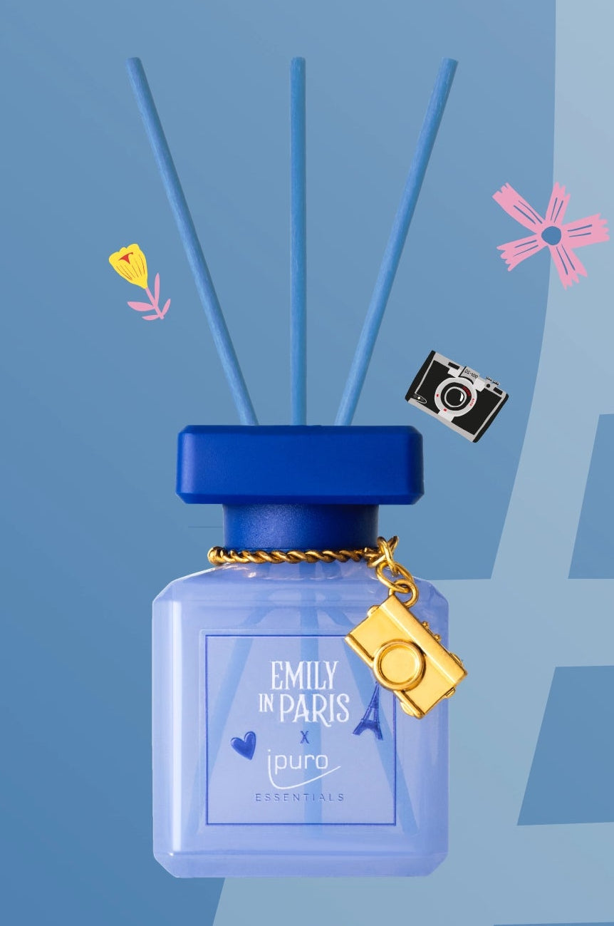 Zwei Parfümzerstäuber in lila und rosa Flaschen mit Emily in Paris-Etiketten, dekorativen Anhängern (eine Kamera auf der blauen Flasche, der Eiffelturm auf der rosa Flasche) und Paris-Symbolen auf einem geteilten blauen und rosa Hintergrund.