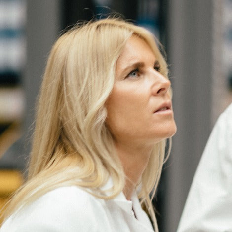 Eine Frau mit langen blonden Haaren, die einen weißen Laborkittel trägt, blickt aufmerksam nach oben. Der Hintergrund ist unscharf und deutet auf ein Labor oder eine wissenschaftliche Umgebung hin.