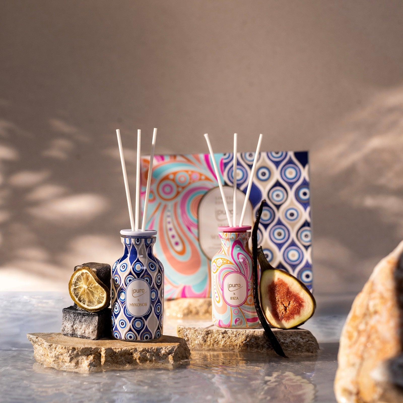 Das LIMITED EDITION Geschenk-Set Ibiza & Mykonos von ipuro zeigt blaue und rosafarbene Deko-Diffusoren auf Steinplatten mit Akzenten von getrockneten Limetten und Feigen, begleitet von einer farbenfrohen Box vor neutralem Hintergrund.