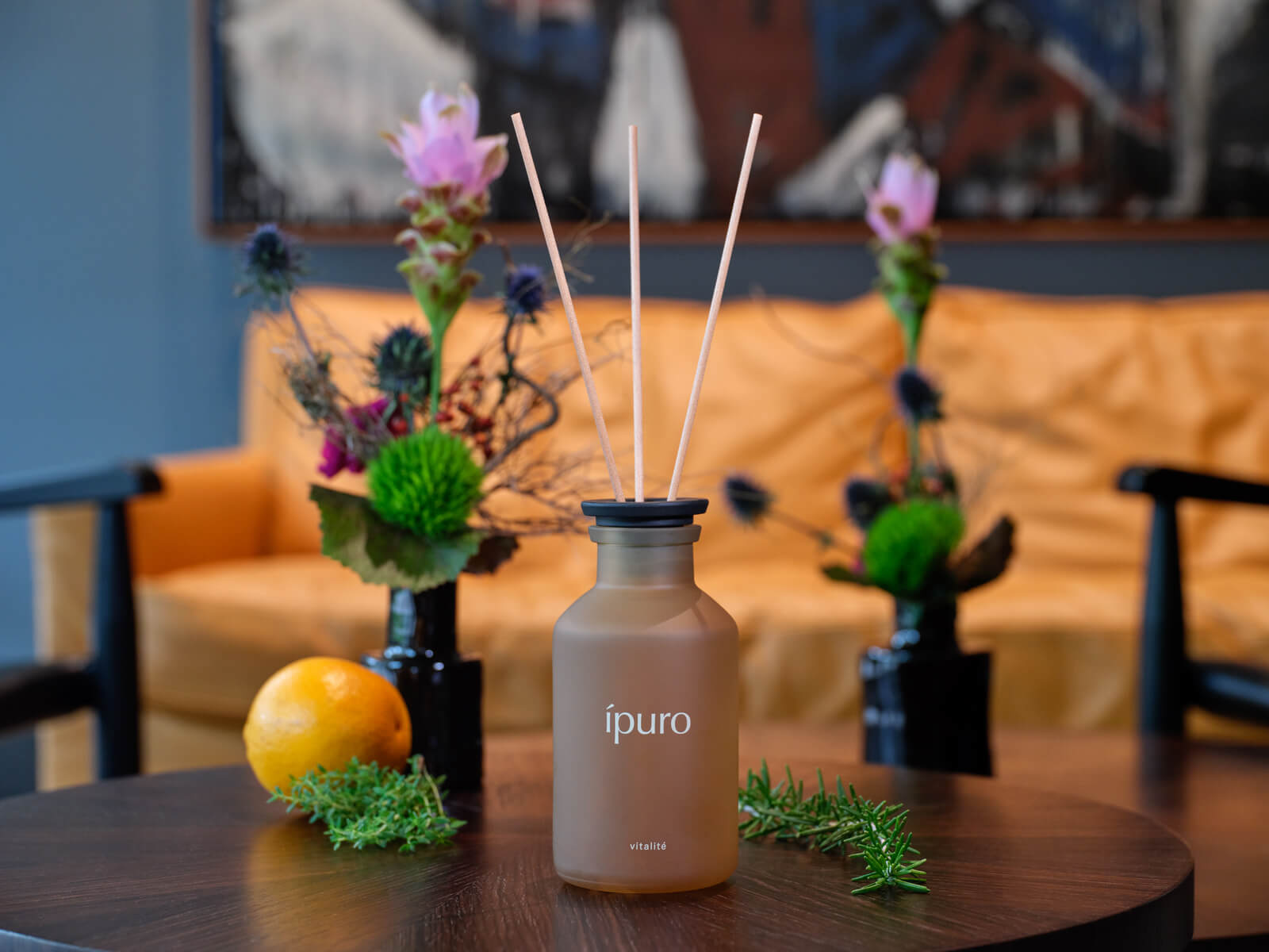 Eine Diffusorflasche aus Milchglas mit der Aufschrift ípuro und Schilfrohrstäbchen steht auf einem Holztisch, umgeben von Kräutern und einer Orange. Im Hintergrund stehen zwei Blumenarrangements und eine gelbe Couch.
