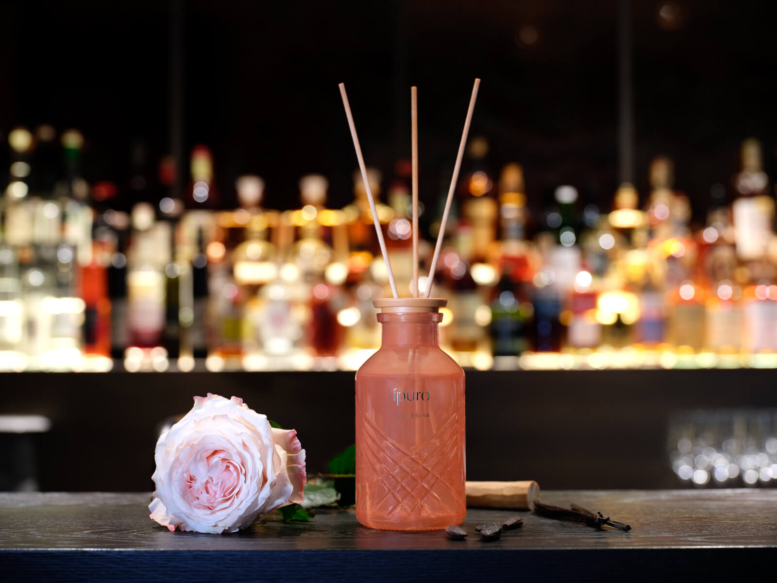 Eine rosafarbene Glasflasche mit hölzernen Schilfrohren steht auf einer dunklen Fläche neben einer blassrosa Rose, mit einer Reihe von verschwommenen Schnapsflaschen im Hintergrund.