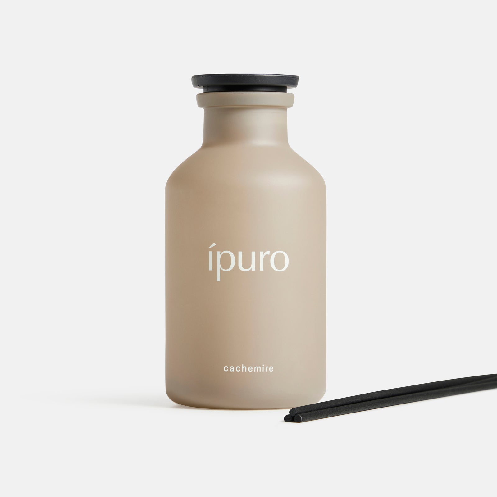Ein mattbeiger Raumduft ipuro cachemire Flakon von ipuro steht aufrecht mit einer schwarzen Kappe neben zwei schwarzen Diffusorstäbchen auf weißem Grund und erinnert an elegante Rose und warme Zedernholznoten.