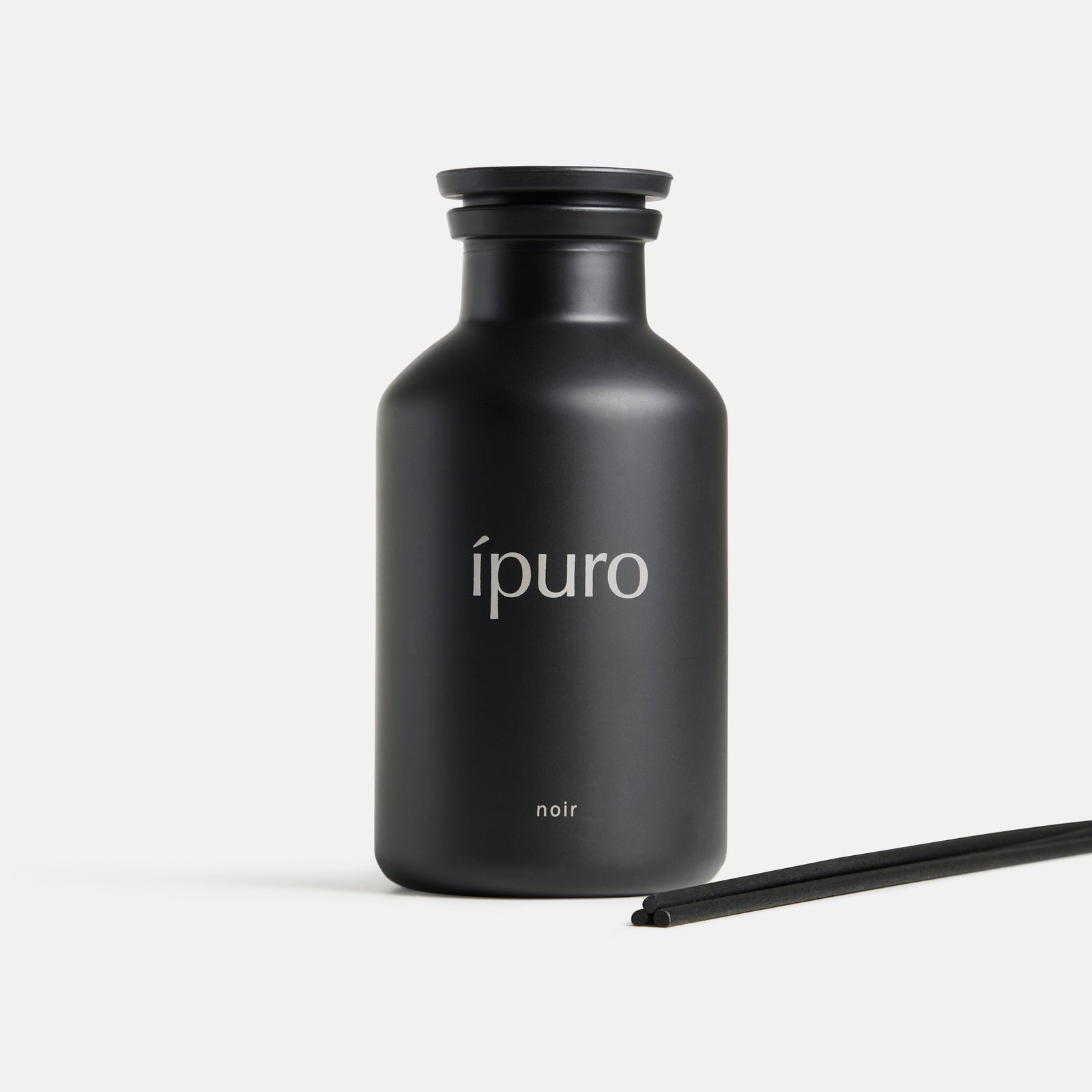 Ein mattschwarzer ipuro-Flakon mit der Aufschrift "Raumduft ipuro noir" steht aufrecht auf weißem Grund und erinnert an Sandelholz und Patchouli, daneben liegen zwei schwarze Diffusorblätter.