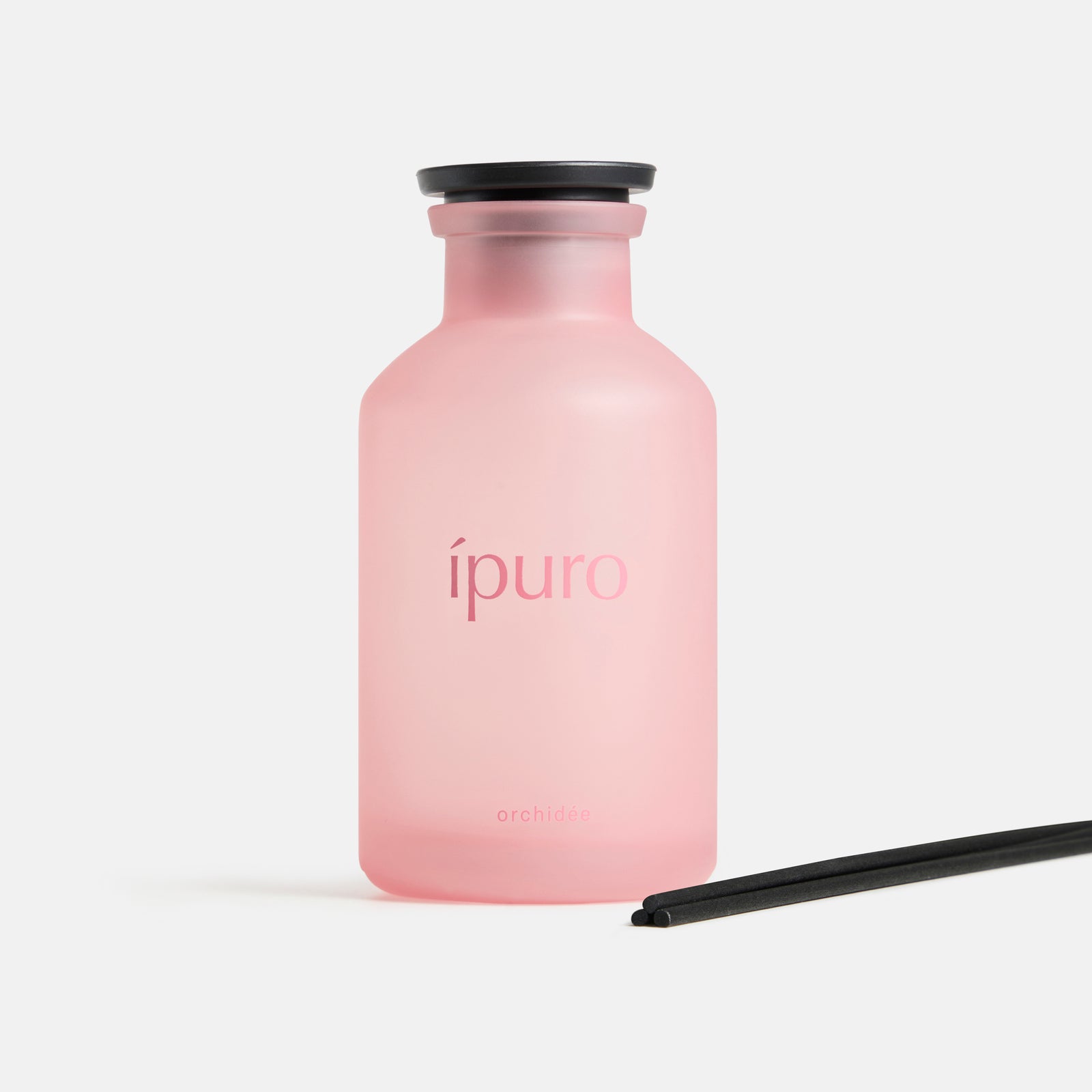 Ein mattrosafarbener ipuro-Glasflakon mit der Aufschrift "Raumduft ipuro orchidée" mit einer schwarzen Kappe und zwei schräg davor platzierten schwarzen Diffusorstäbchen vor einem schlichten weißen Hintergrund - das ist der charakteristische Duft von ipuro.