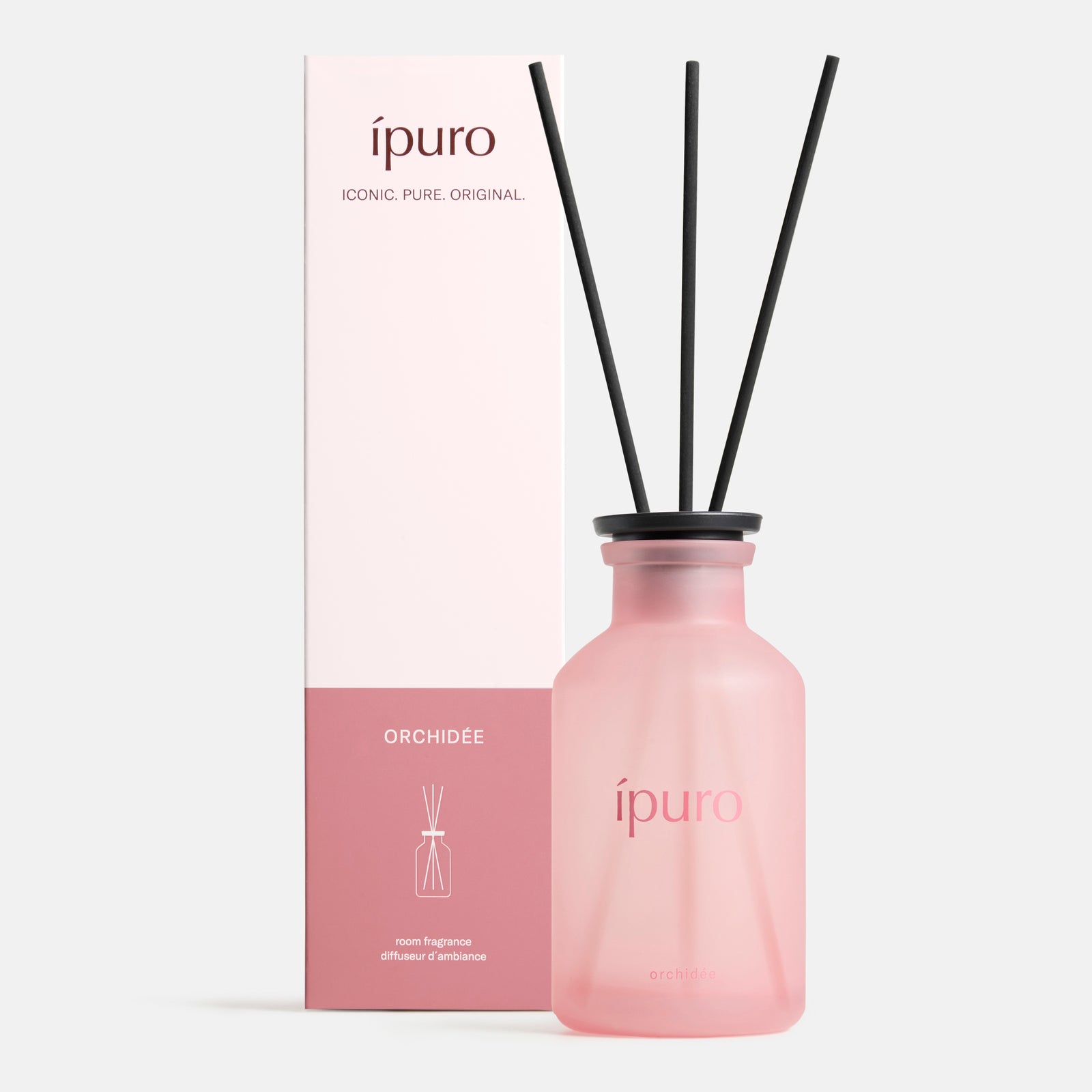 Ein rosafarbener Milchglasflakon mit der Aufschrift ipuro Raumduft orchidée und schwarzen Diffusorstäbchen steht neben einer passenden rosa-weißen Schachtel von ipuro, die zarte Noten von Veilchen und sizilianischer Limone hervorruft.
