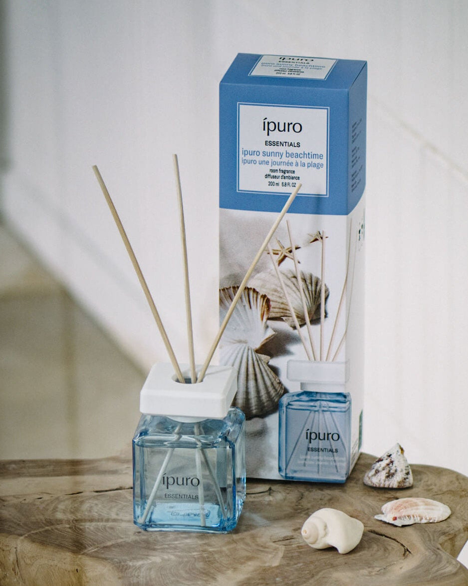 Ein quadratischer Glasflakon des ipuro Sunny Beachtime Schilfdiffusors mit Holzstäbchen steht auf einer Holzfläche neben Muscheln, die blau-weiße Verpackung mit Muschelmotiven im Hintergrund.